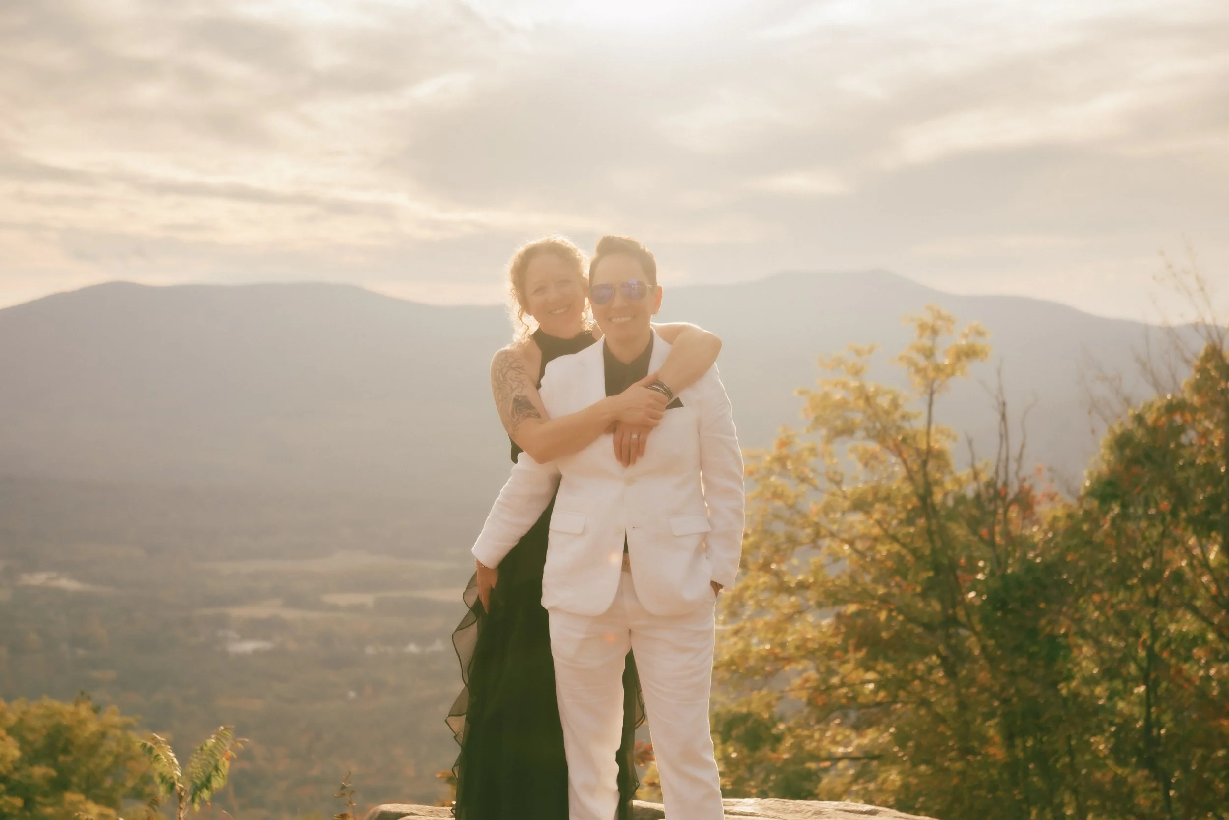 Two people, one wearing a white suit and sunglasses and the other in a black dress, standing outdoors with mountains and trees in the background during sunset, smiling and hugging.