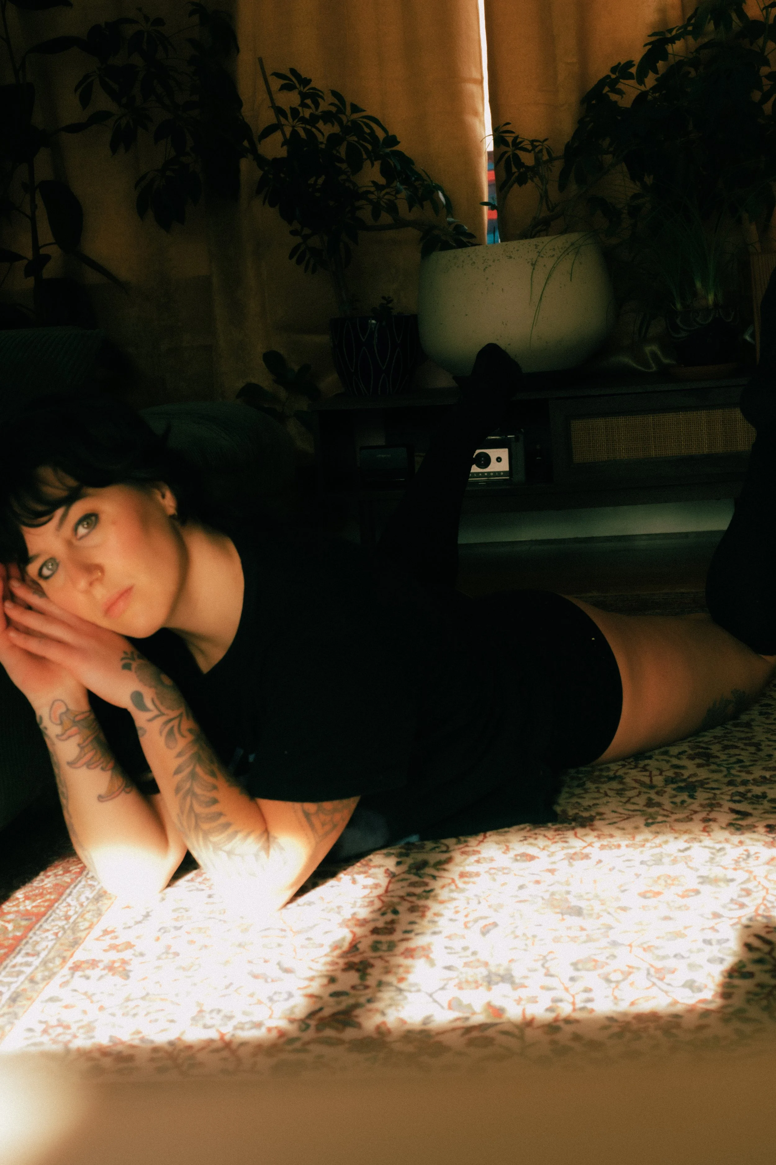 A woman with tattoos lying on her side on a patterned rug, supporting her head with her hand, in a dimly lit room with plants and curtains in the background.