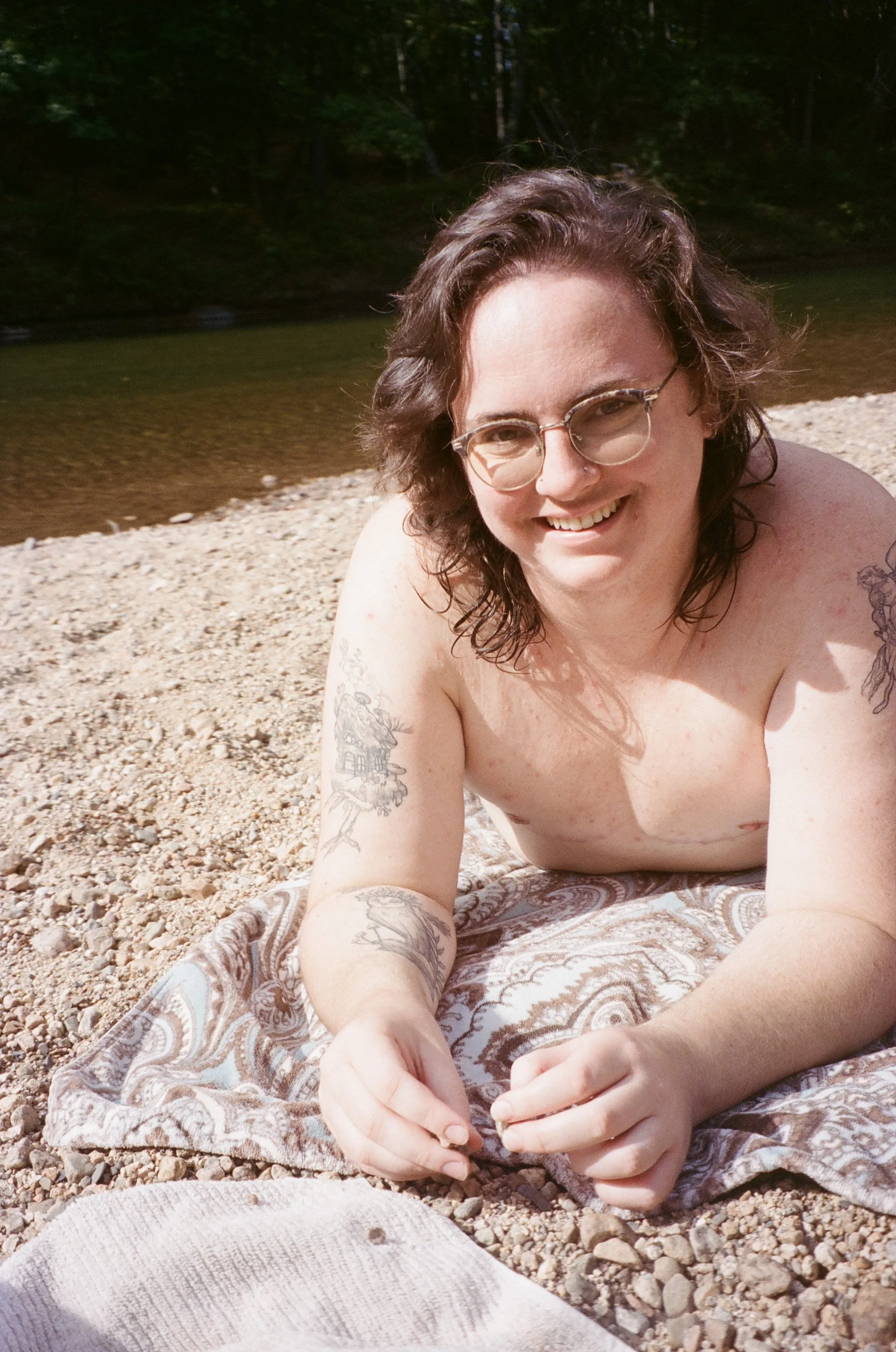 A person with glasses, shoulder-length hair, and tattoos lying on a patterned blanket on a pebbled riverbank, smiling at the camera.