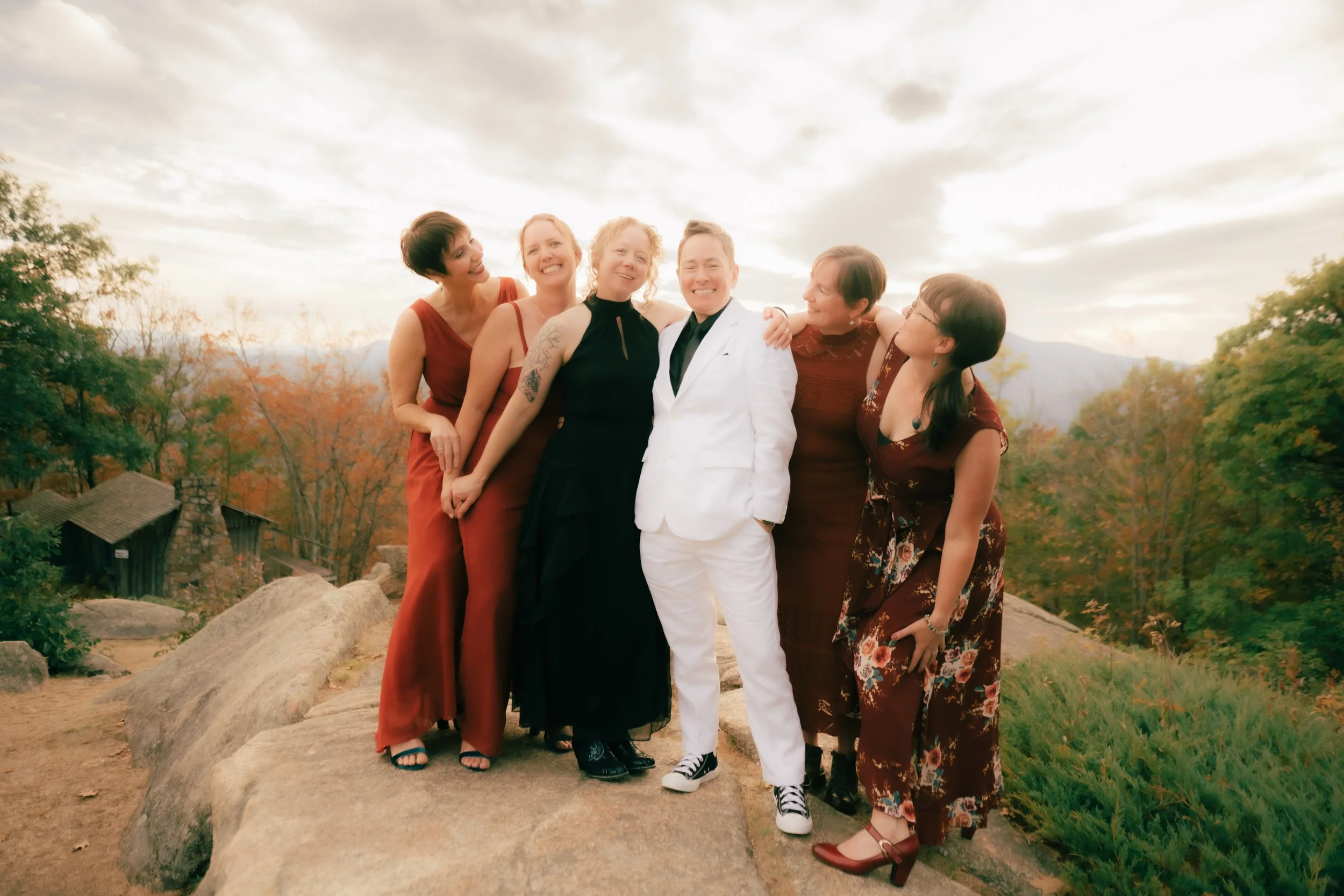 A group of six people, five women and one man, standing on rocks outdoors with trees in autumn foliage behind them. The man is dressed in a white suit, and the women are wearing dresses of various colors, smiling and enjoying each other's company.