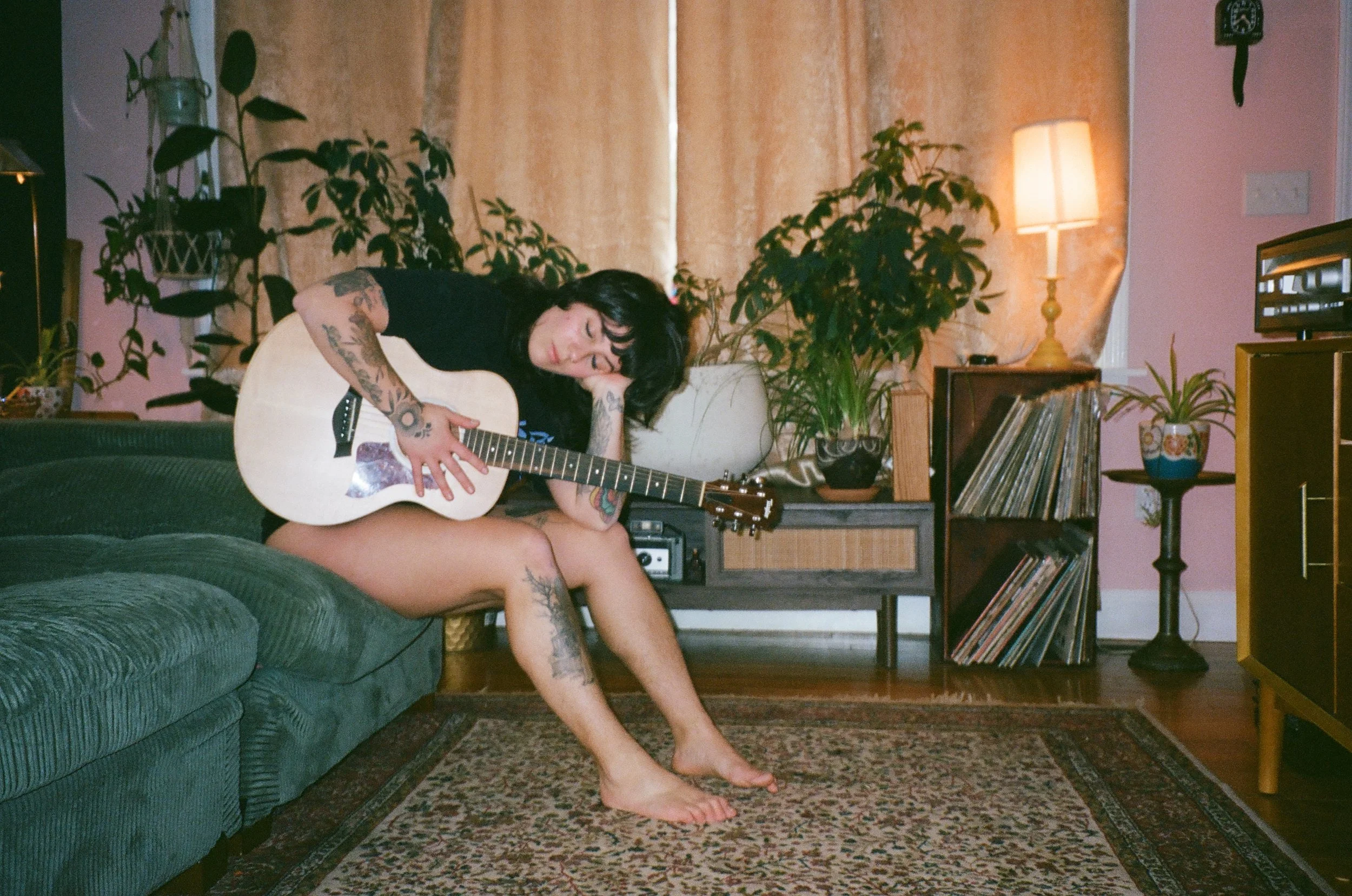 A young woman with tattoos sits on a sofa in a cozy living room, playing an acoustic guitar with her eyes closed, resting her head on her hand, surrounded by houseplants, a lamp, and vinyl records.