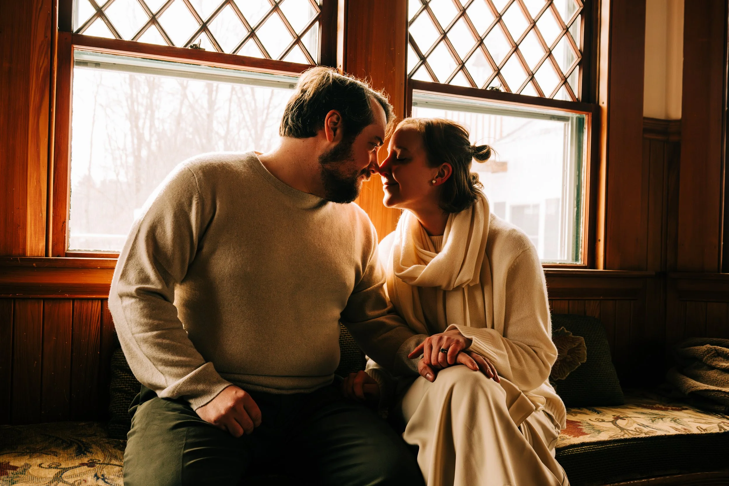 A couple sitting close together indoors, touching foreheads and holding hands, by a window with wooden framing and diamond-shaped glass panels.