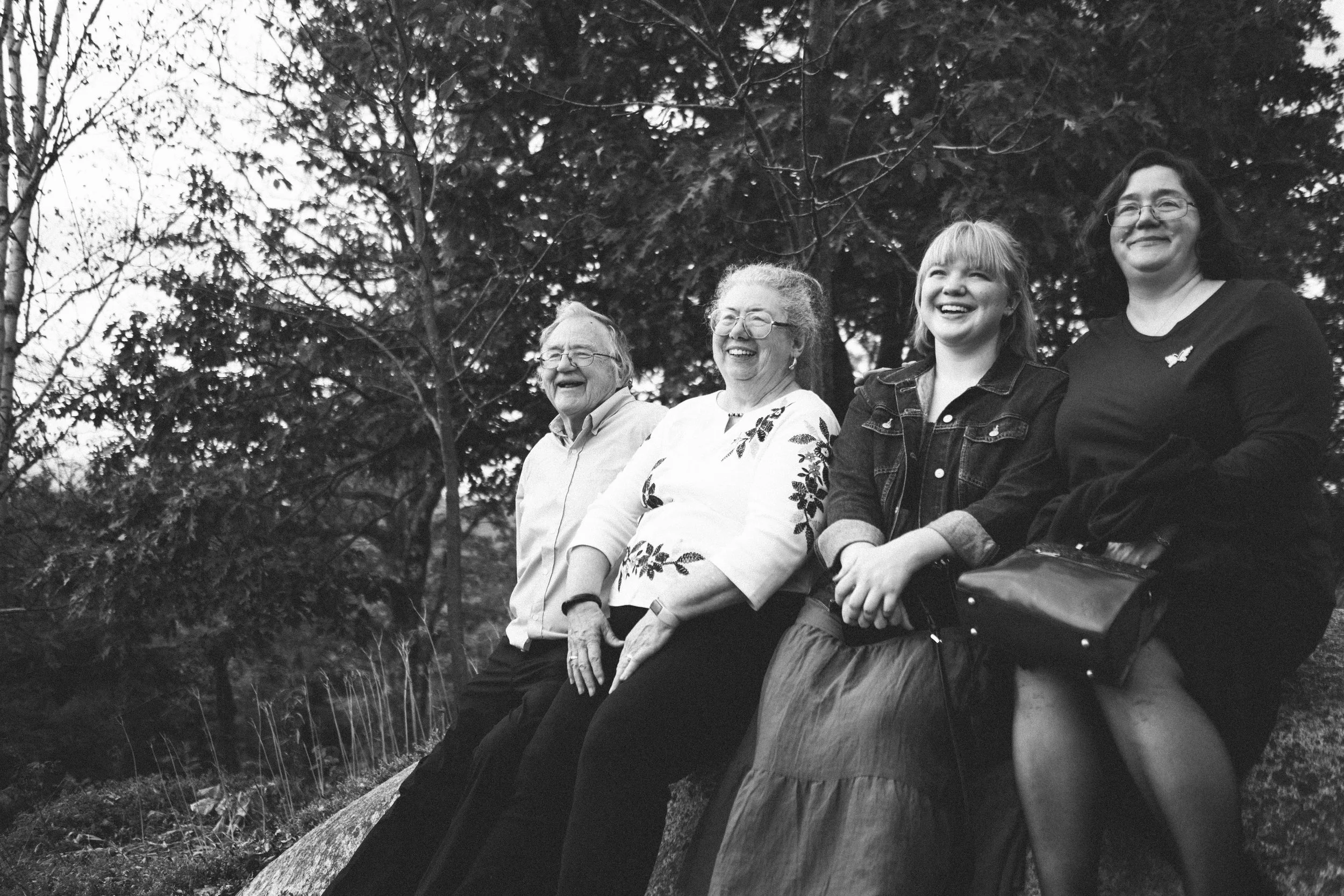 Four women sitting outdoors on a log, smiling and laughing, surrounded by trees.
