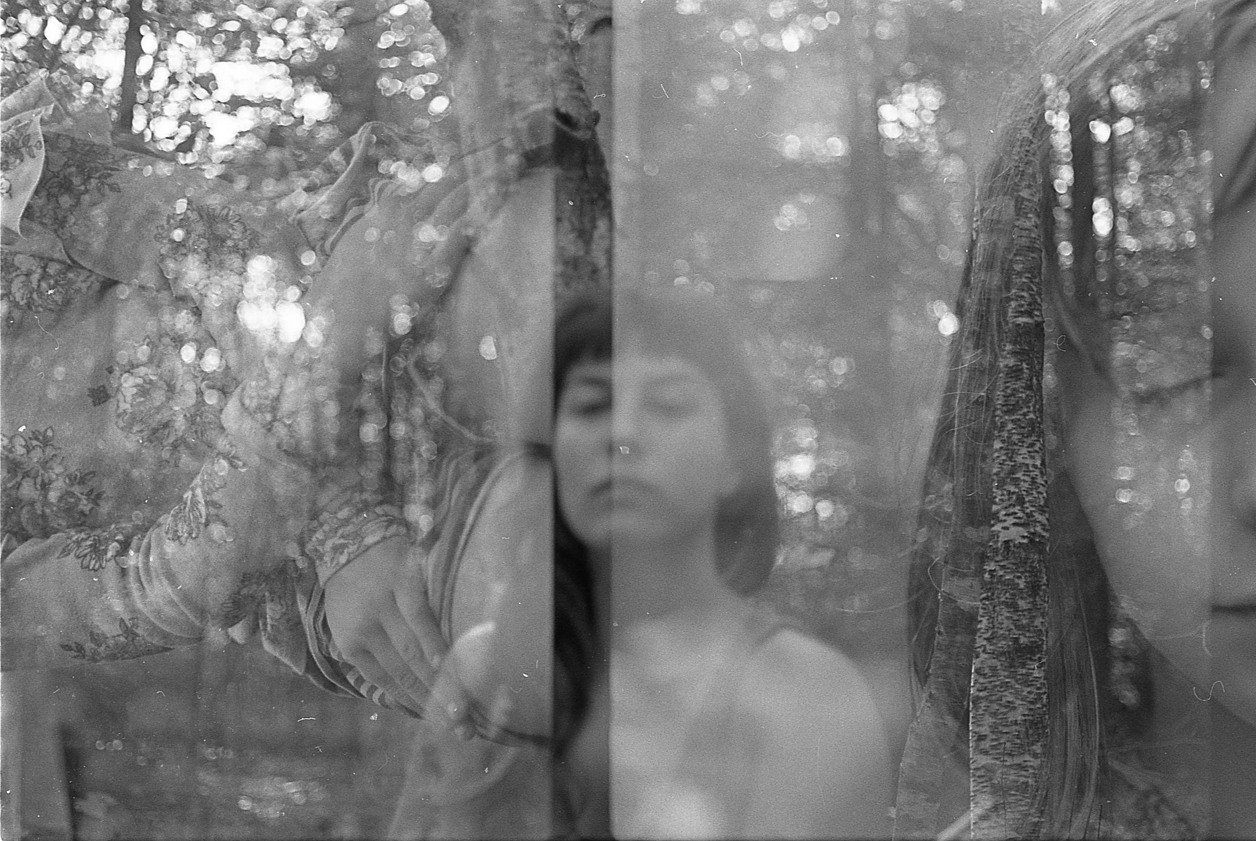 A young woman with long hair leaning against a tree, with her eyes closed and a serene expression, surrounded by dense trees and dappled sunlight.