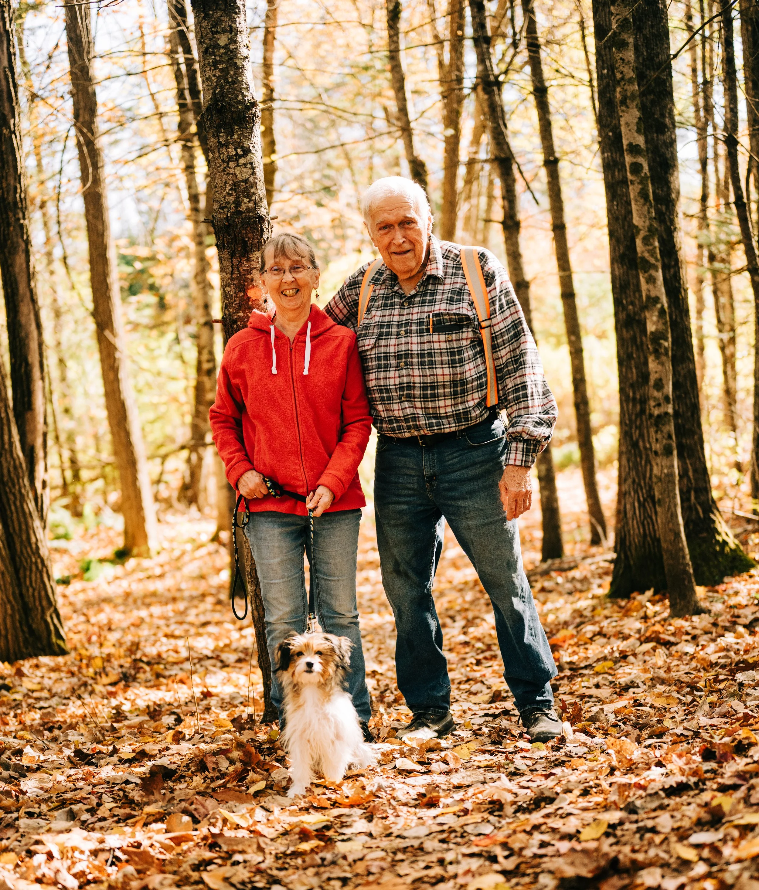 An elderly couple hiking in a forest with their small dog, surrounded by fallen autumn leaves.