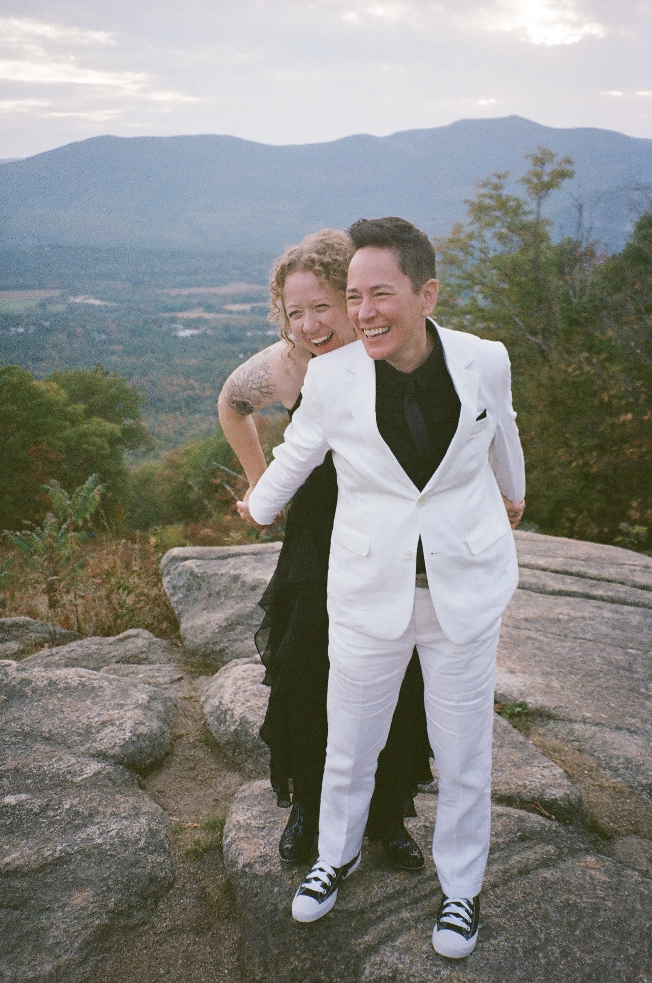 A couple laughing and standing on rocks outdoors with mountains and trees in the background. The woman has curly blonde hair and a tattoo on her arm, wearing a black dress. The man has short dark hair, wearing a white tuxedo jacket, black shirt, and 