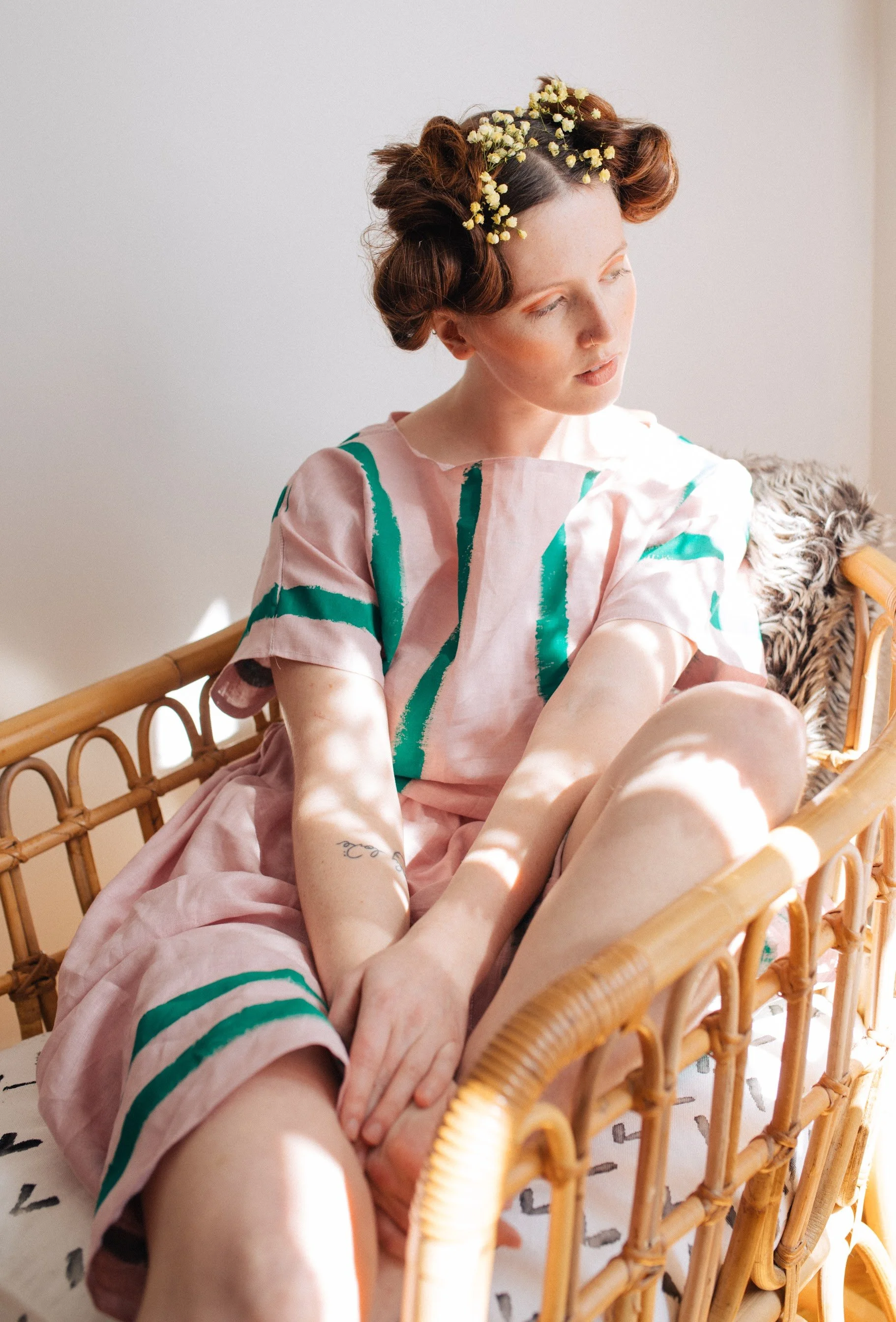 A woman with styled hair and a floral headband sitting on a wicker bench, wearing a light pink dress with green and dark pink patterns, with her legs crossed and hand resting on her leg, bathed in natural sunlight.