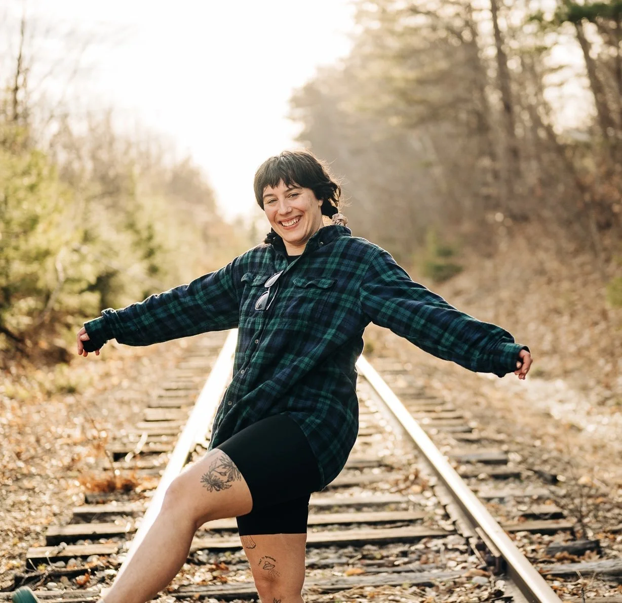 A woman with short dark hair wearing a plaid shirt and black shorts standing on railway tracks surrounded by trees with autumn foliage, smiling with arms outstretched.