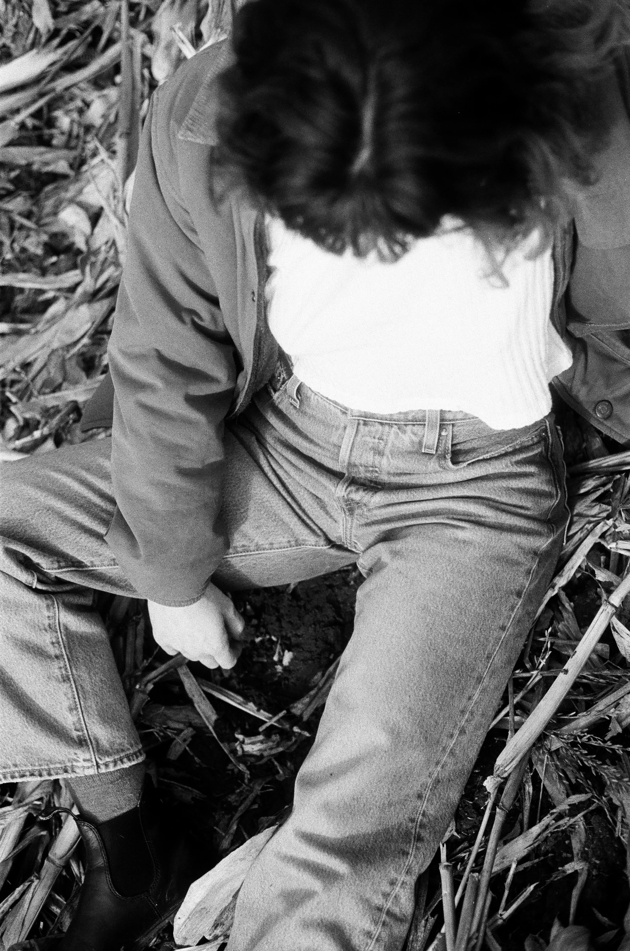 Top view of a person sitting in a field of tall plants or crops, wearing jeans, a jacket, and a shirt.