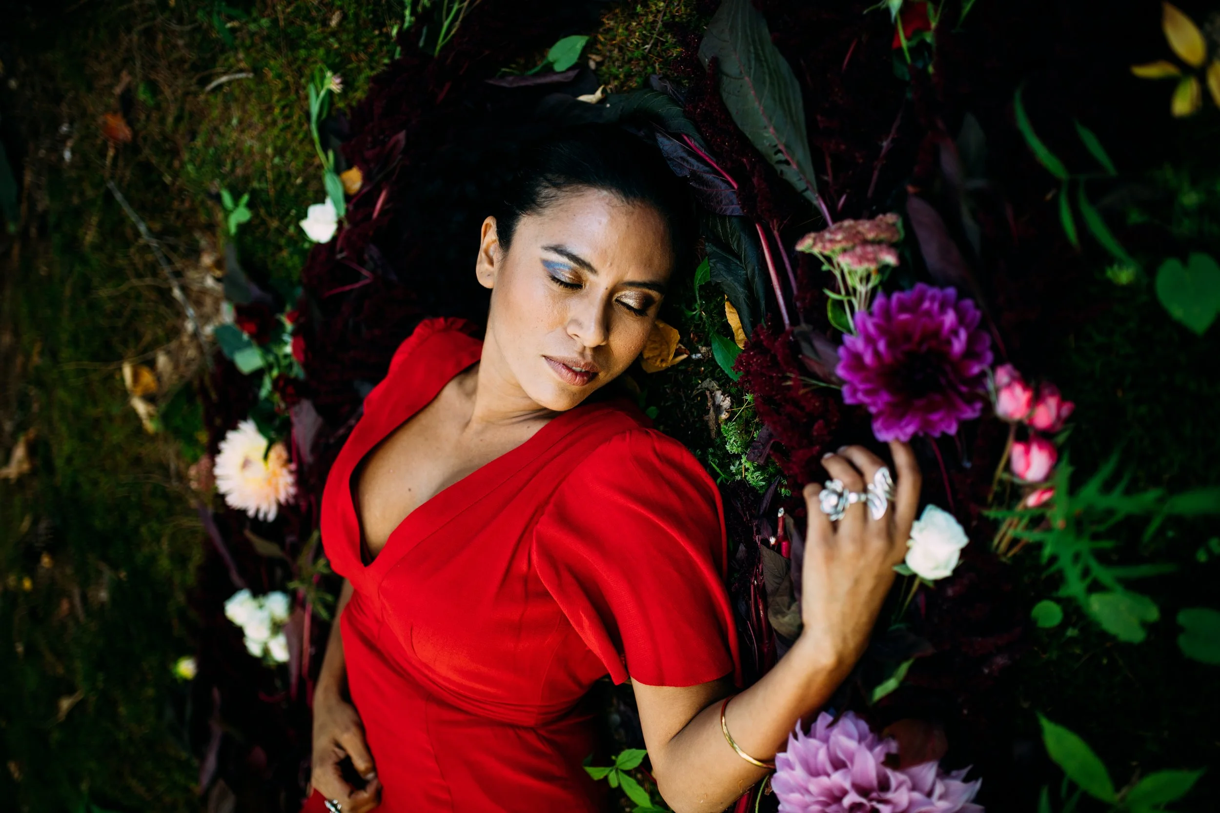 A woman in a red dress lying on the ground surrounded by colorful flowers with her eyes closed.