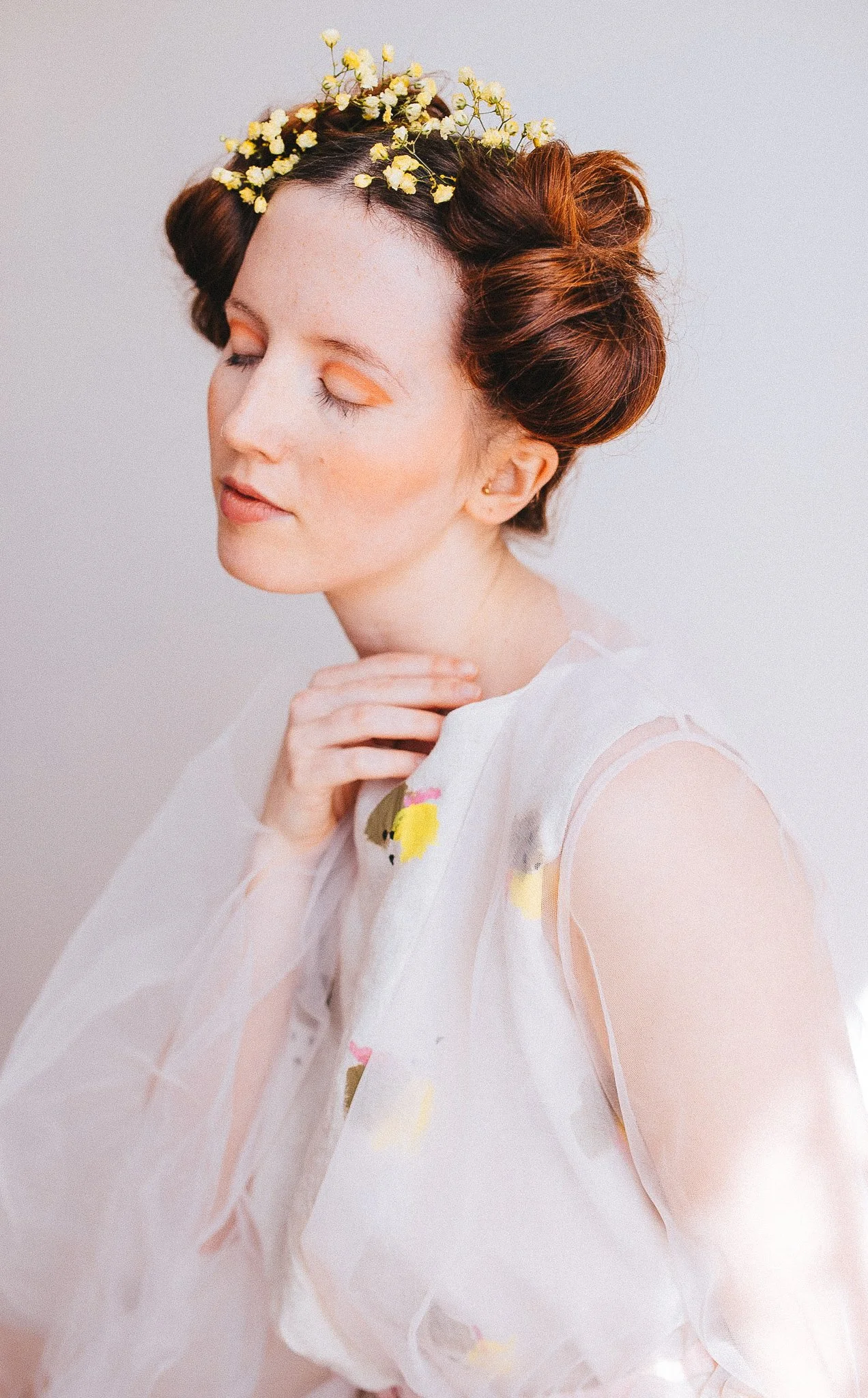 A woman with red hair styled in waves wearing a crown of yellow flowers, soft makeup, and a sheer white dress with embroidered flowers, posed with her eyes closed and hand gently touching her neck.