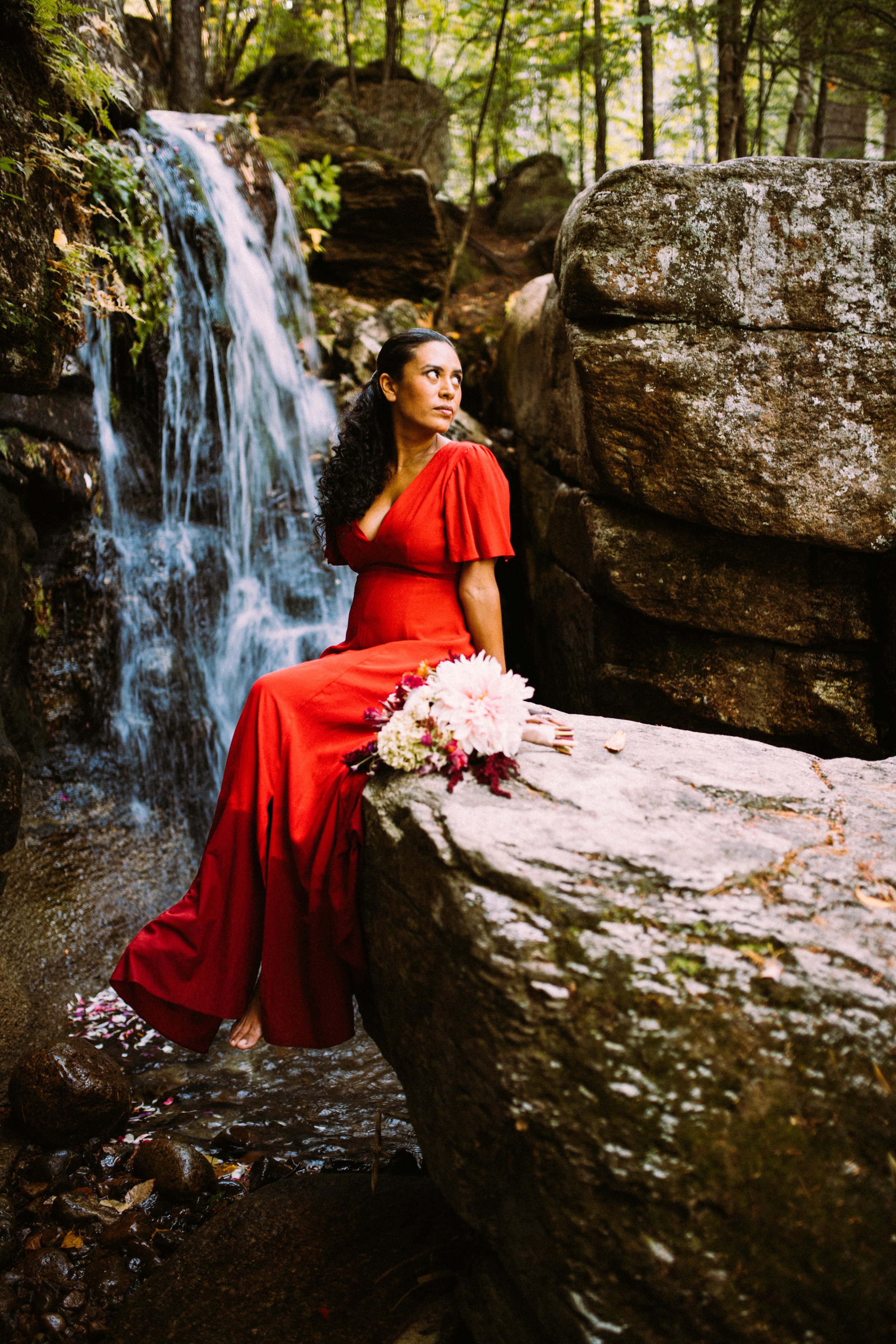 A woman in a red dress sitting on a rock near a waterfall in a forest setting, with a bouquet of flowers placed on the rock beside her.