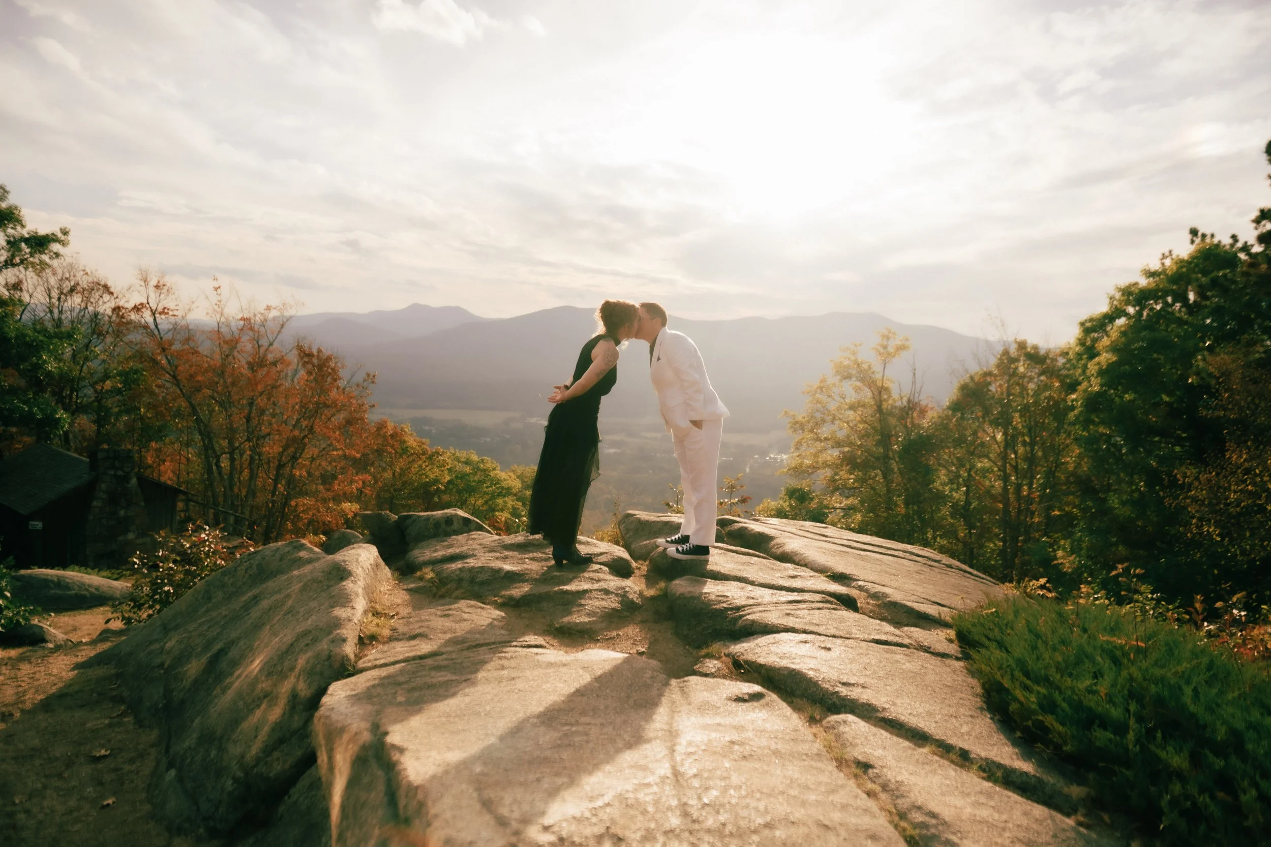 A couple sharing a kiss on a rocky hilltop during sunset, surrounded by autumn trees and mountain scenery.