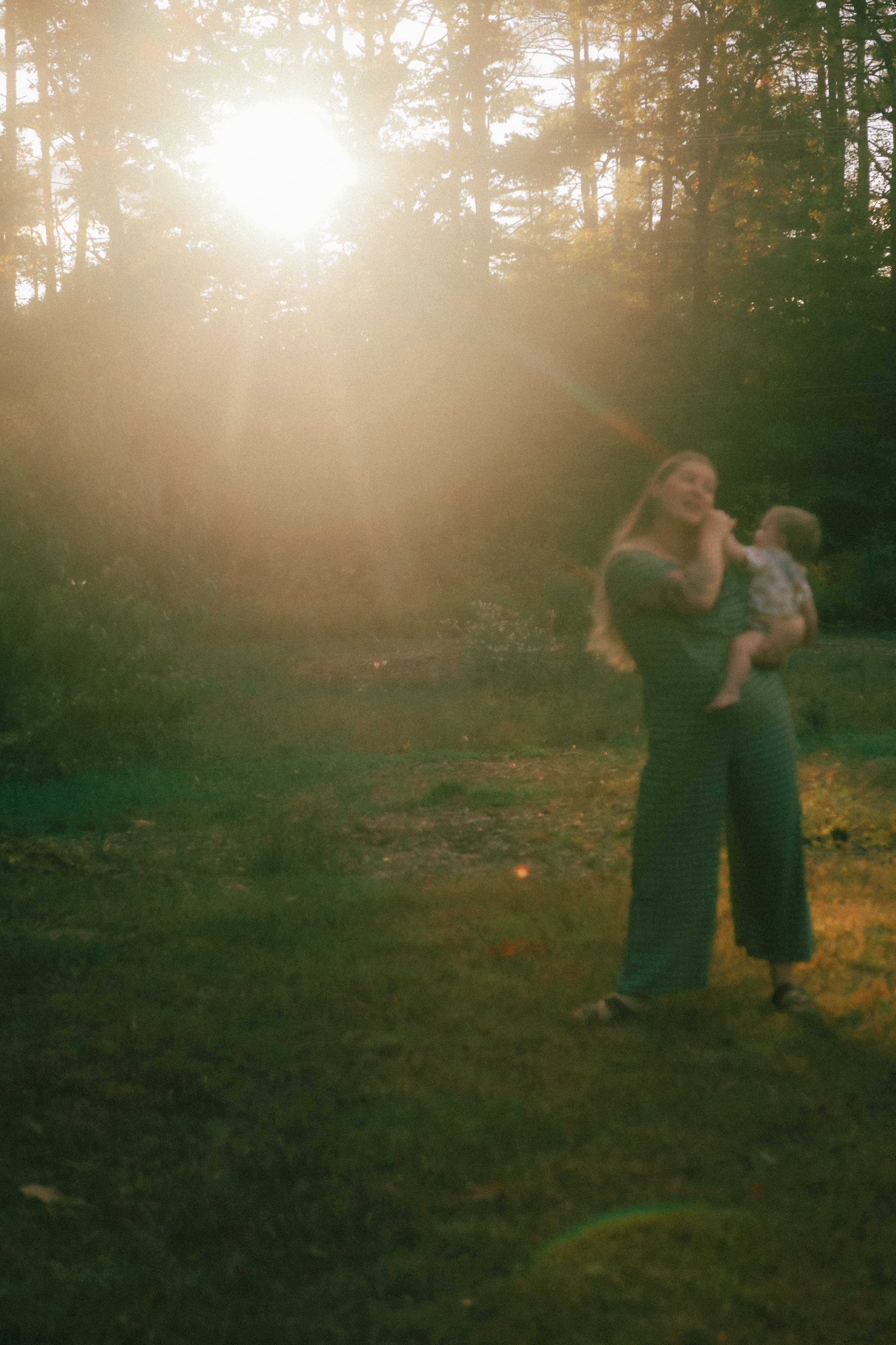 A woman holding a baby outdoors during sunset, with trees and sunlight creating a golden glow.