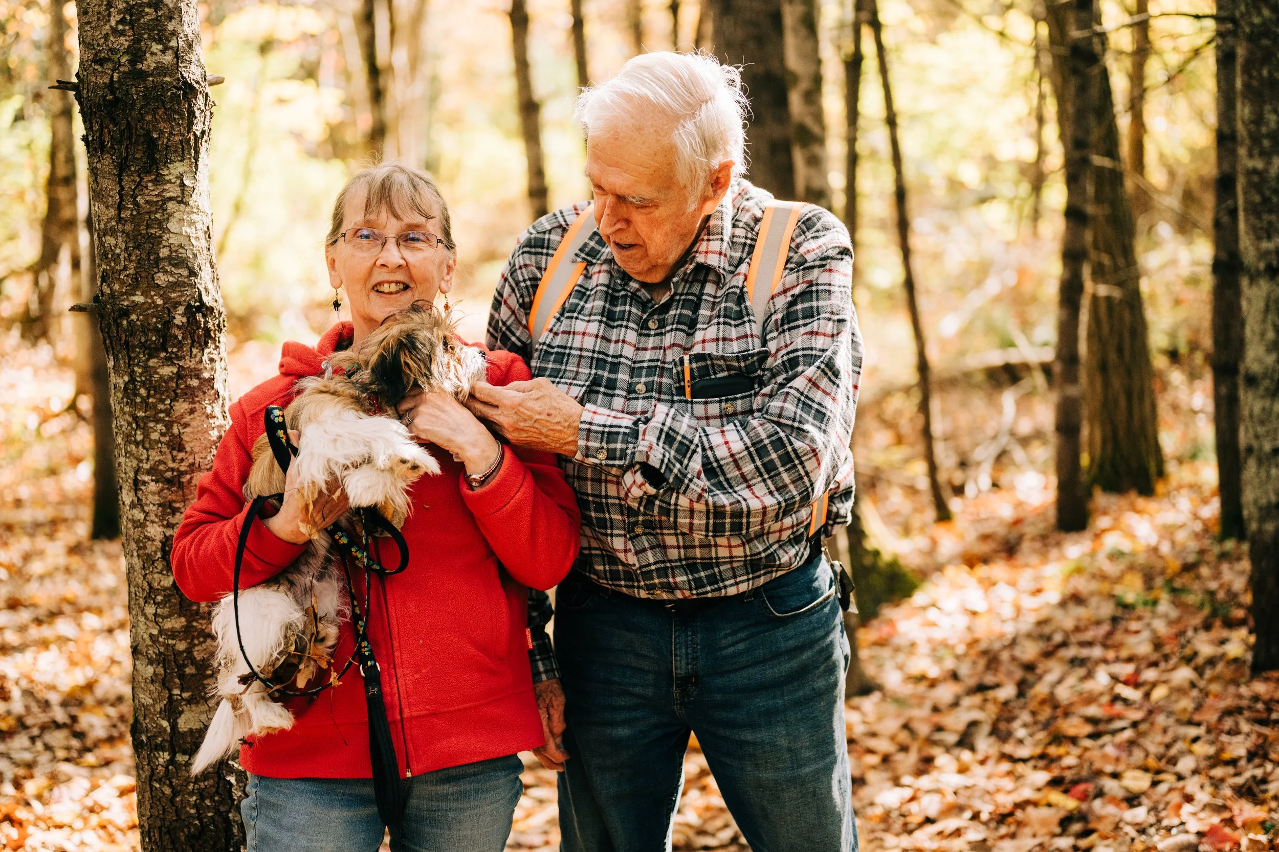 An elderly couple smiling and playing with their dog in a forest during autumn.