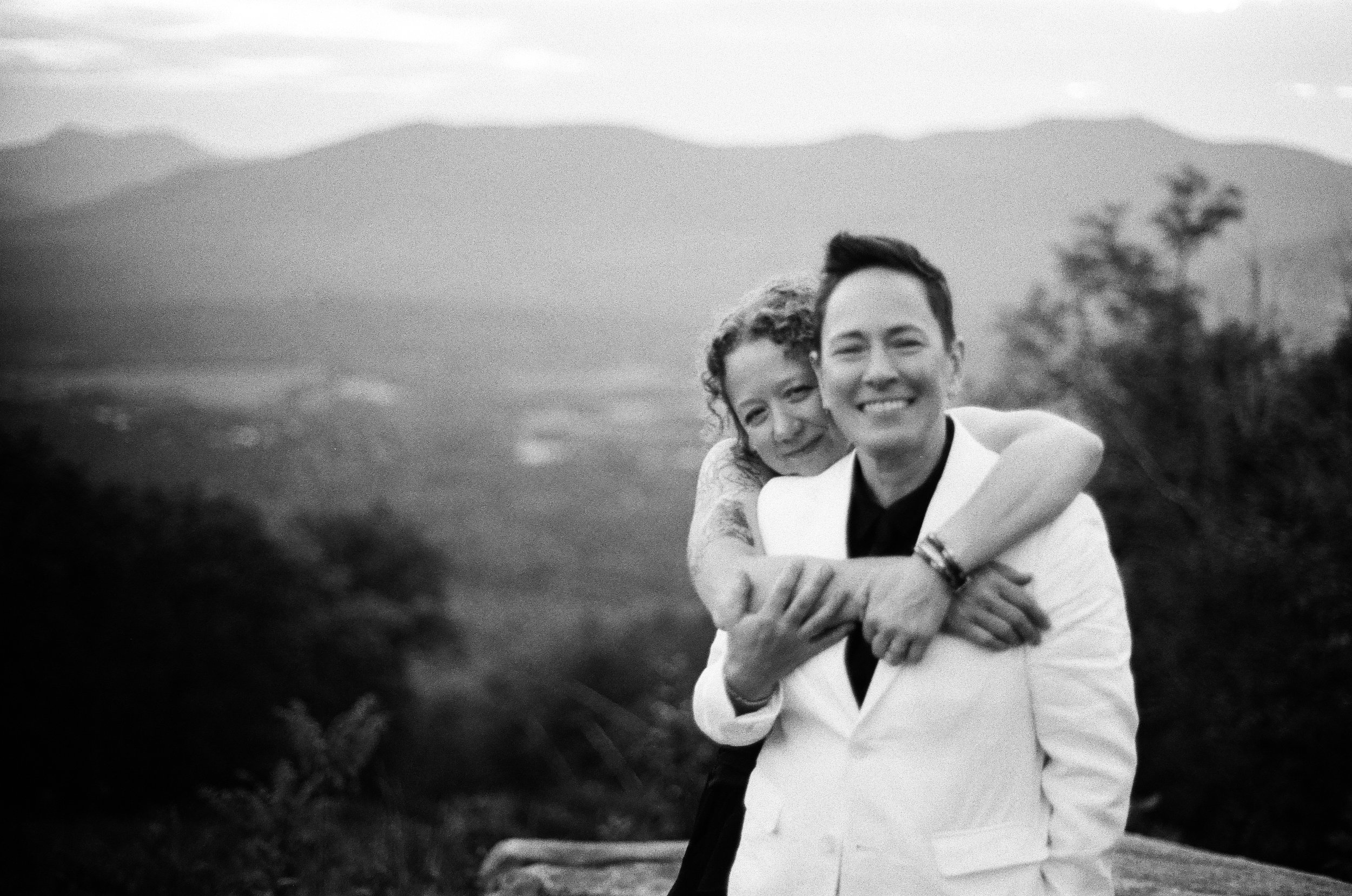 A smiling couple, the woman with curly hair and the man with short dark hair, embracing outdoors with mountain scenery in the background, black and white photograph.