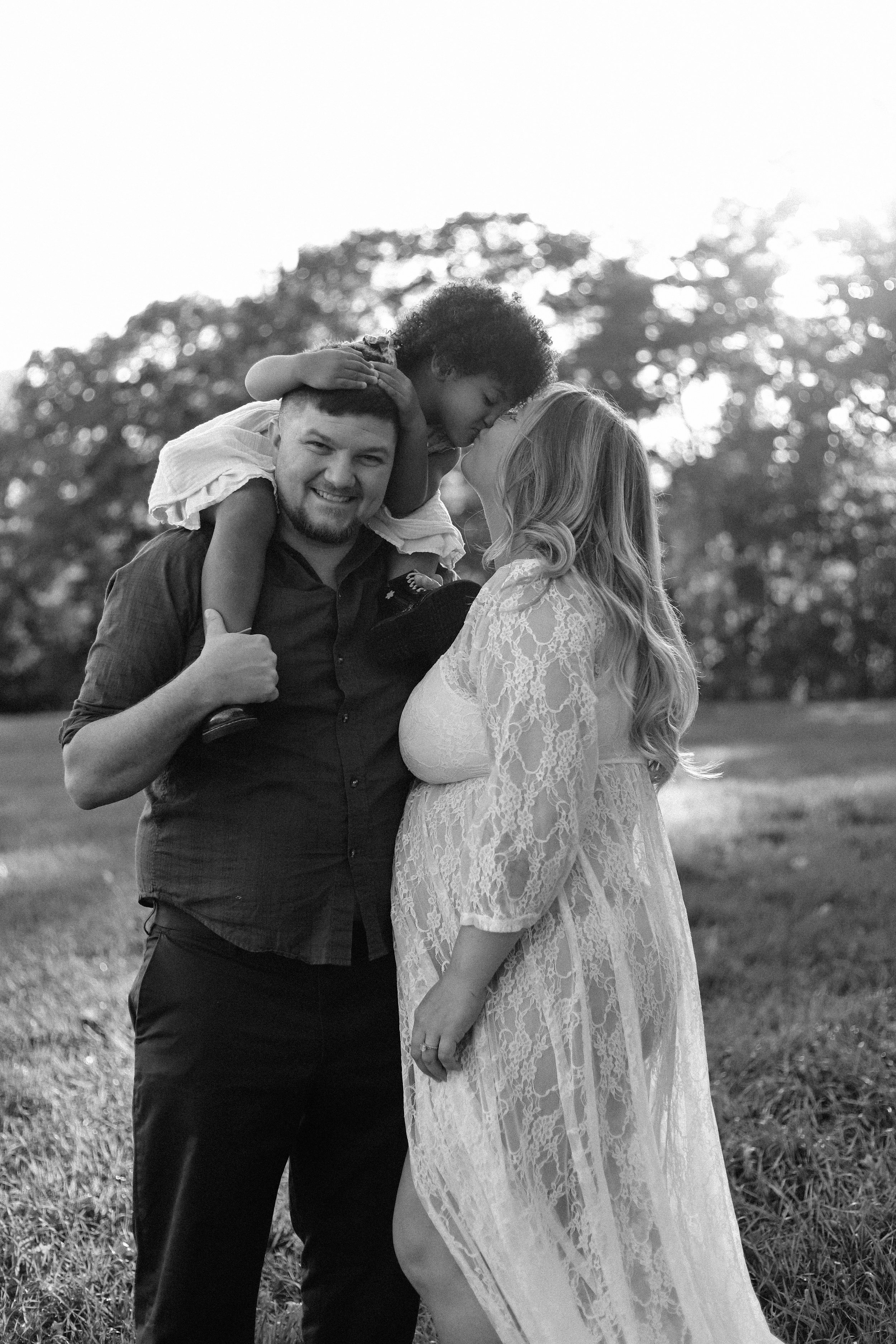 A black and white photograph of a smiling family outdoors. A man carries a little girl on his shoulder, and a woman stands next to them in a lacy dress, with her face close to the girl. Trees and bright sunlight are in the background.