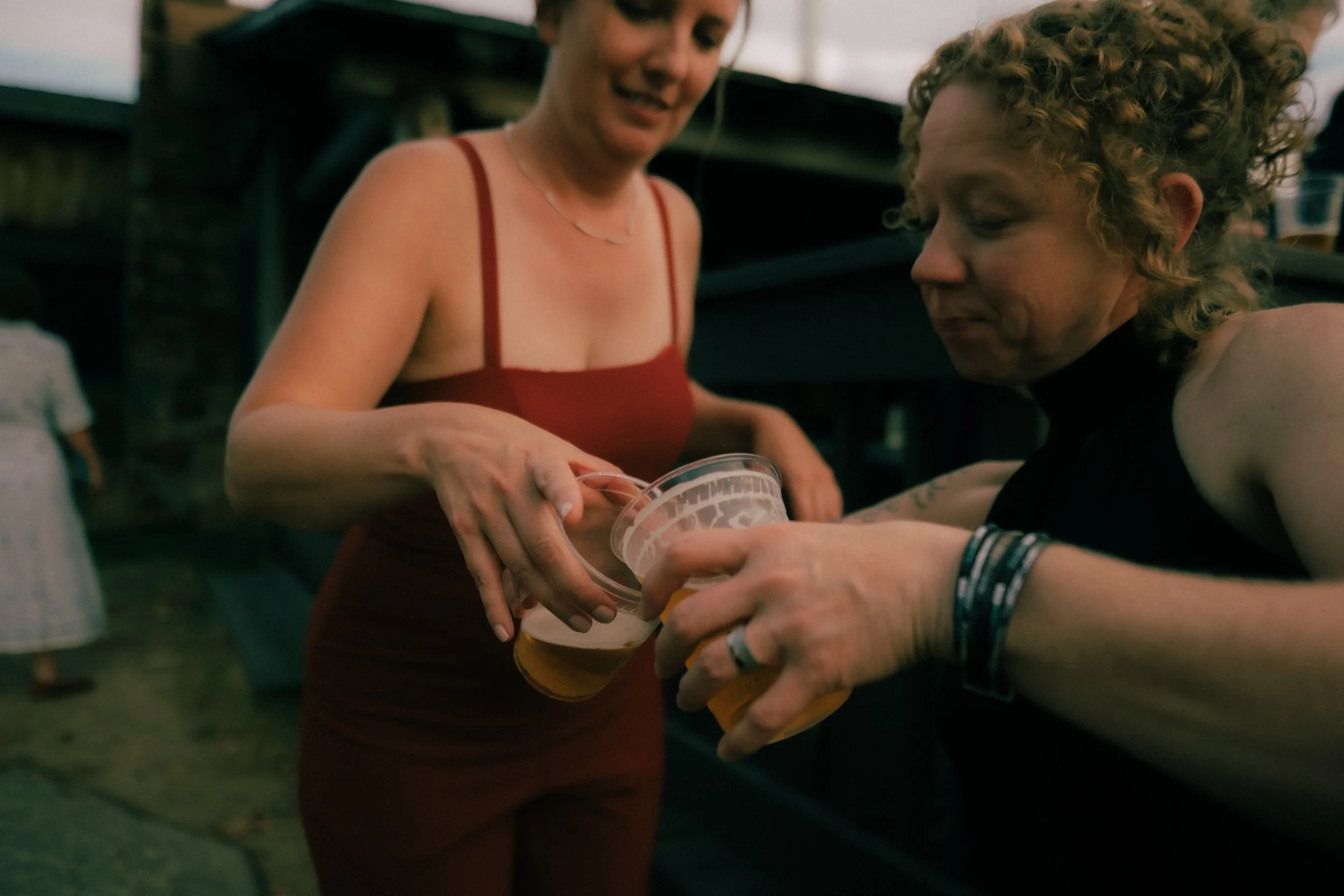 Two women sharing drinks at an outdoor gathering, one woman is handing a drink to the other.