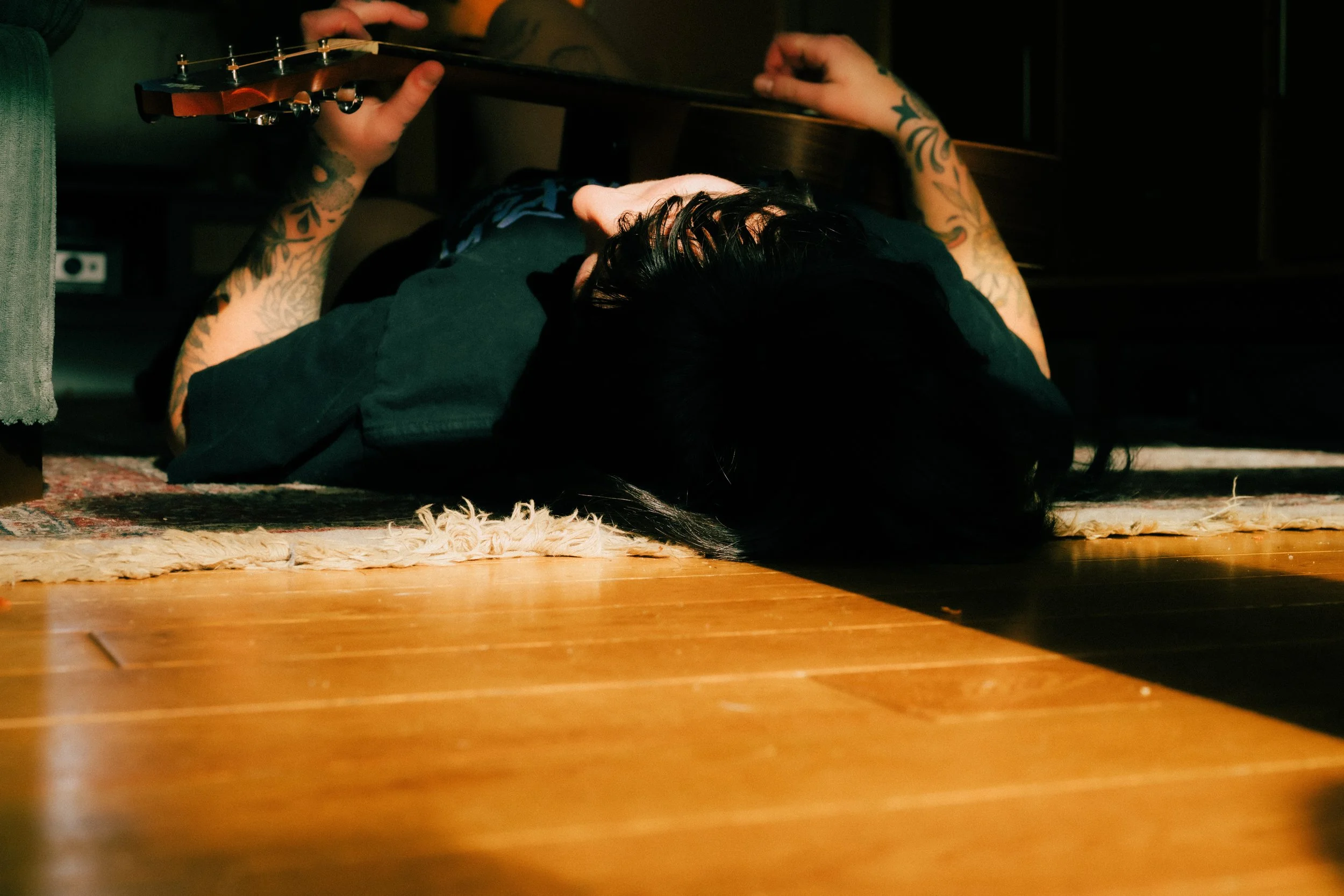 A person lying on their back on a wooden floor, holding an acoustic guitar above their head. They have dark hair, tattoos on their arms, and are wearing dark clothing. The person is resting their head on a small shaggy rug near the edge of the floor,