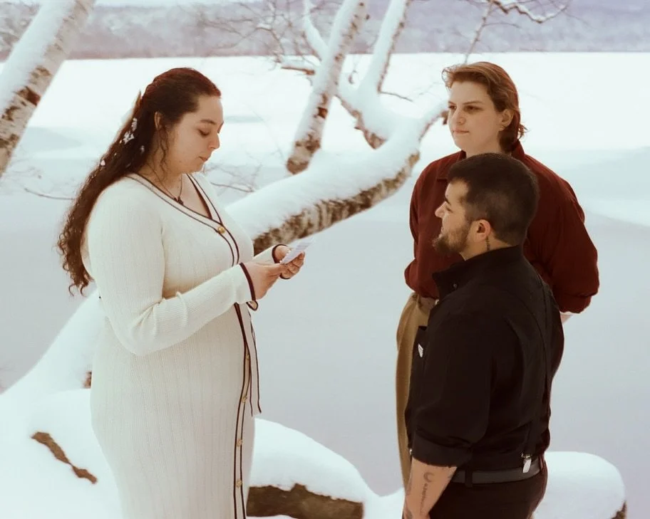 A woman in a white dress reads a paper while two men listen outdoors in a snowy landscape with snow-covered trees.