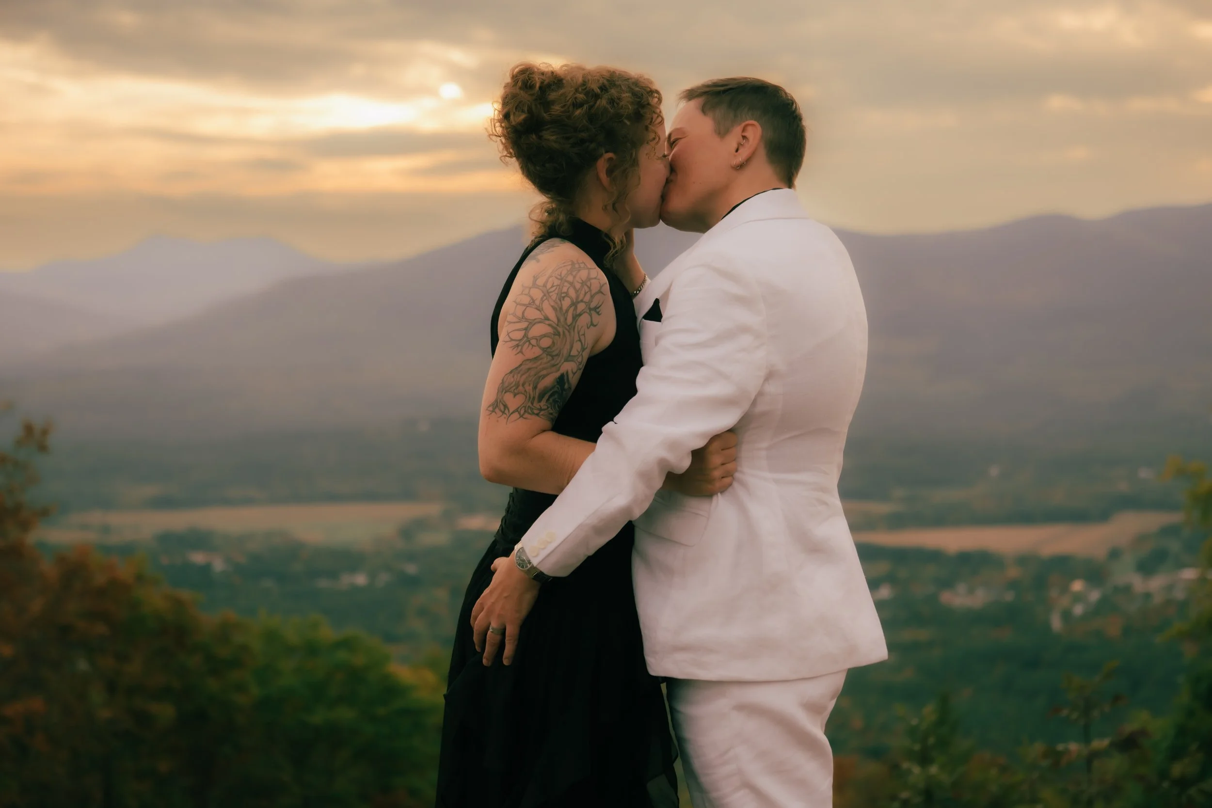 Two women kissing outdoors during sunset, with mountains and trees in the background. One woman wears a black dress and has a tattoo on her arm; the other wears a white suit.