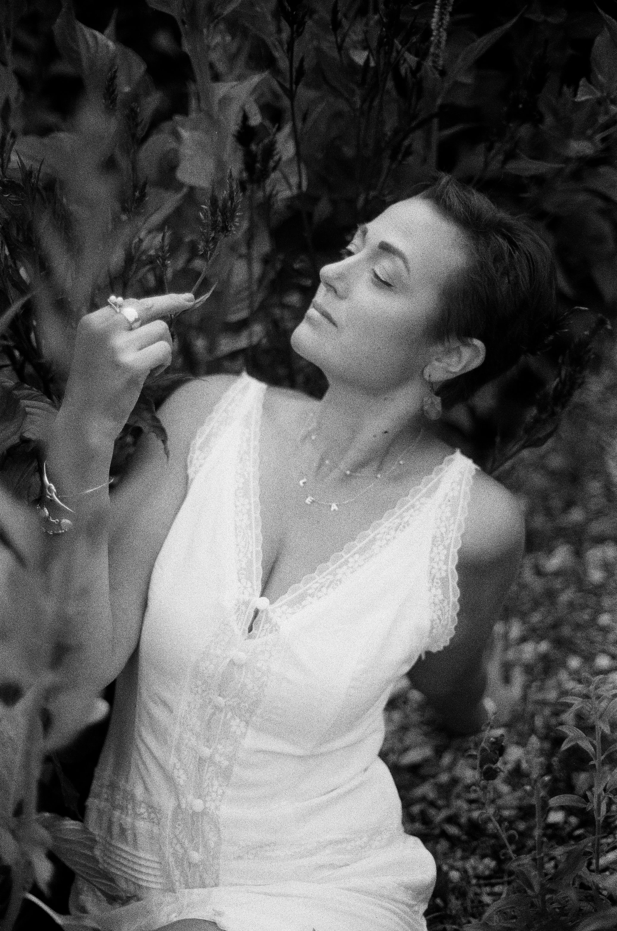 A woman in a light-colored lace dress with her eyes closed, surrounded by plants, holding a small flower near her face in a serene outdoor setting.
