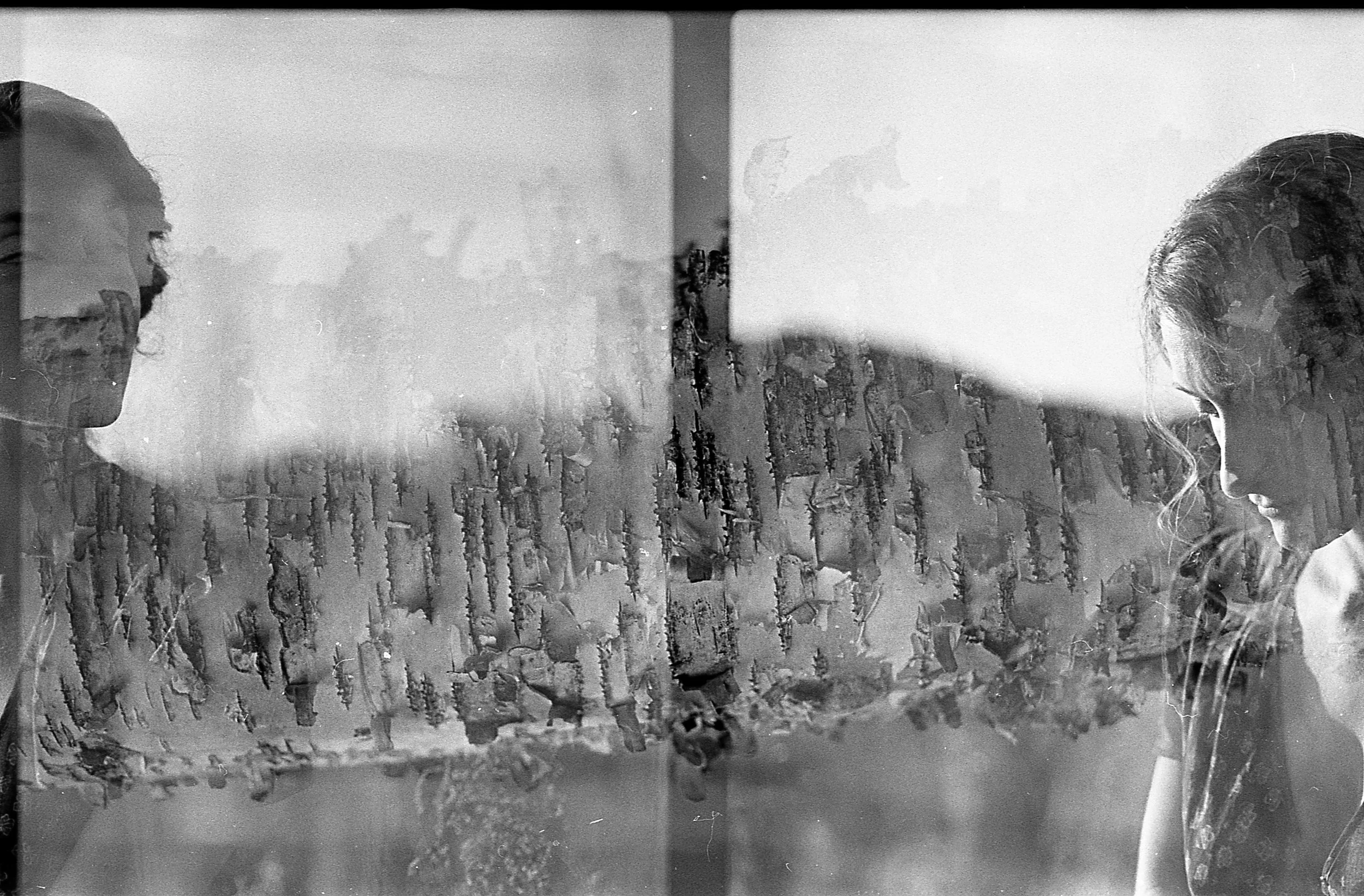 Reflected image of two women with their faces turned downward, seen through a rain-streaked glass window, with an urban cityscape in the background.