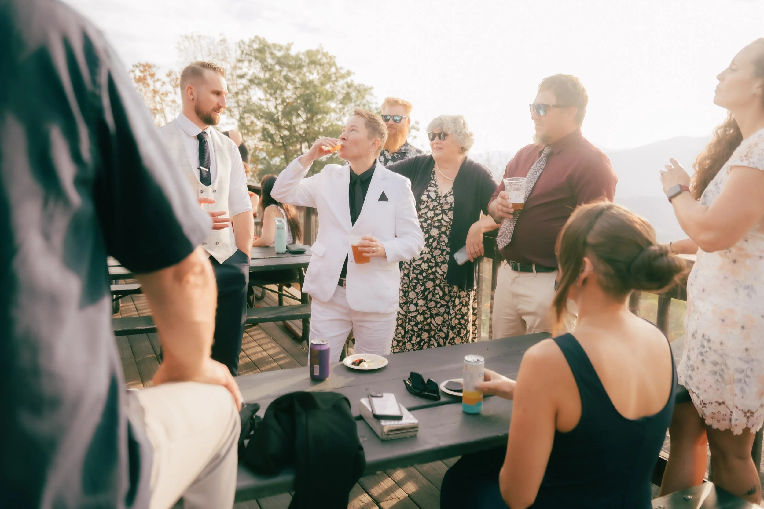 Group of people socializing outdoors on a deck, with some holding drinks, and one person in a white suit eating food.