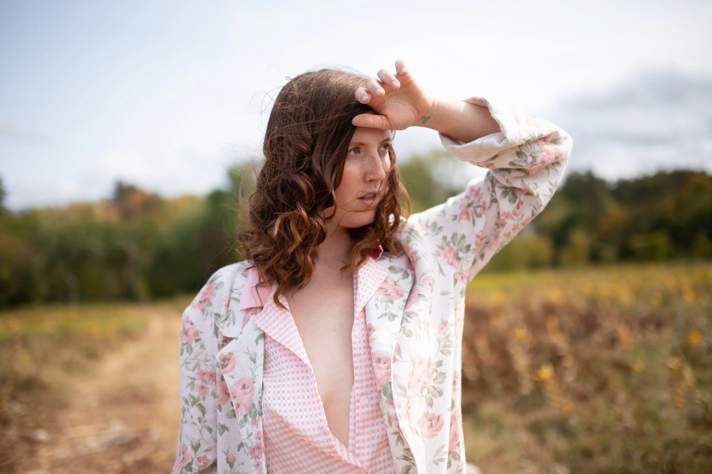 A woman with curly reddish-brown hair stands outdoors in a field, wearing a floral and pink checkered shirt, with her right hand on her forehead, looking off into the distance.