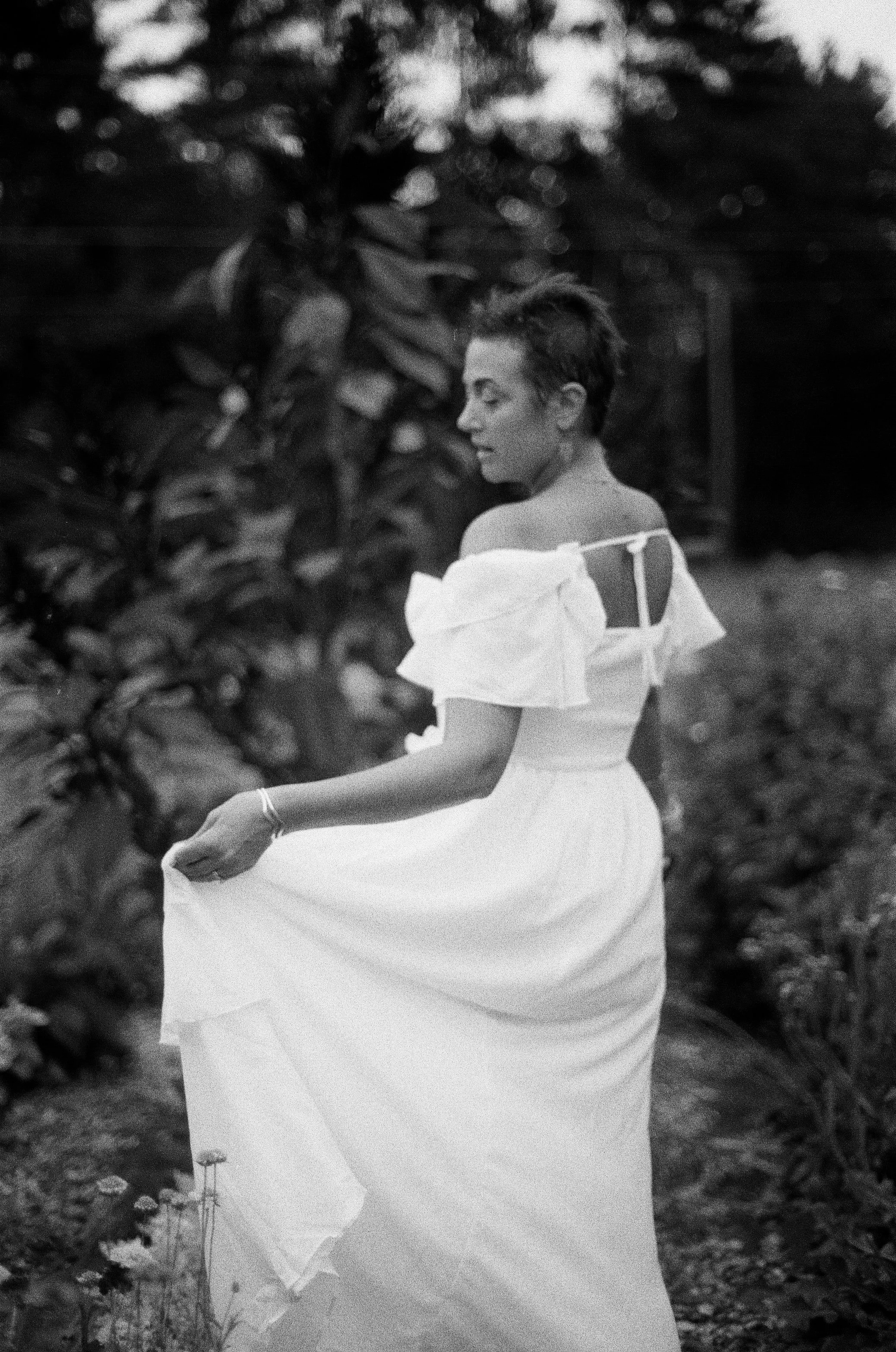 A woman in a white dress walking through a garden, holding the hem of her dress, with trees and foliage in the background.
