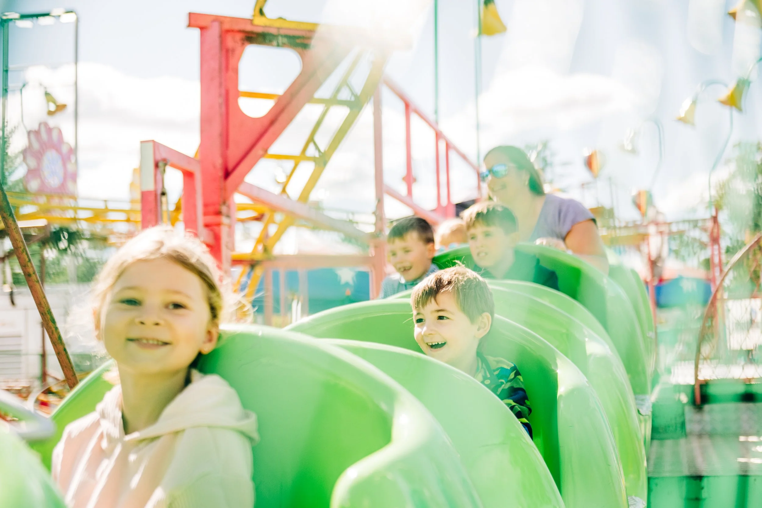Children riding a green roller coaster at an amusement park, with a woman supervising, under a bright sky with clouds.