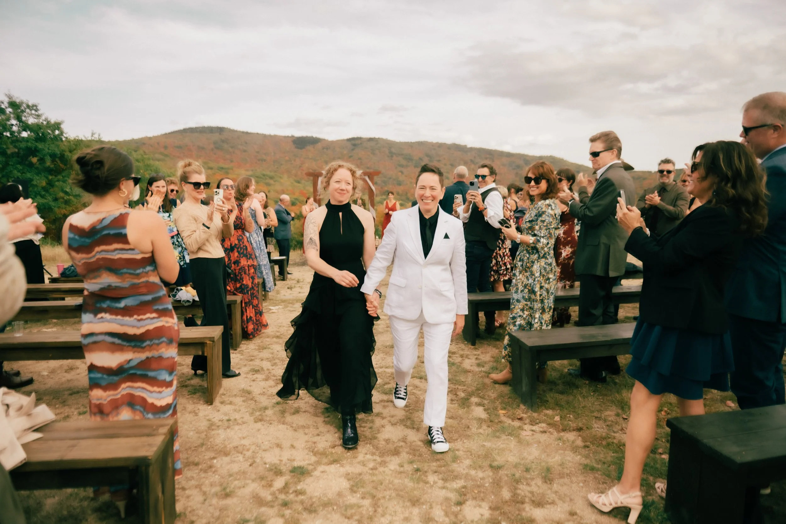 Two women holding hands walking down an aisle at an outdoor wedding ceremony with guests on either side taking photos, with mountains and cloudy sky in the background.