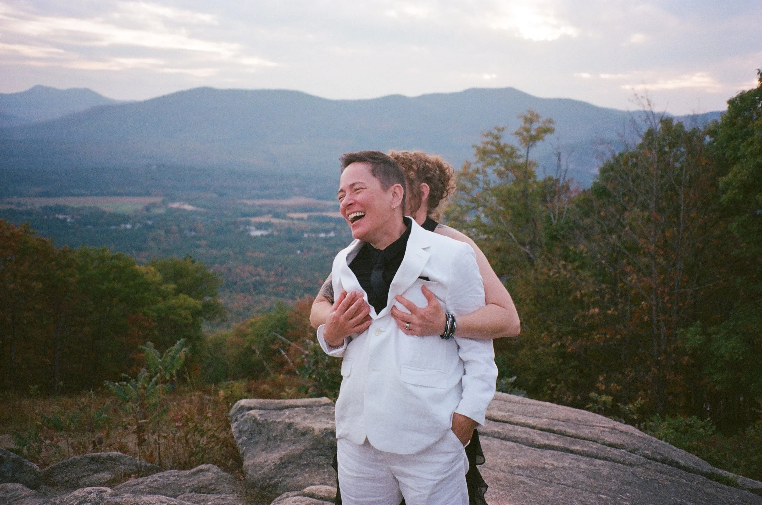 A person in a white suit laughing with someone behind them, in an outdoor mountain setting with trees and rocks.