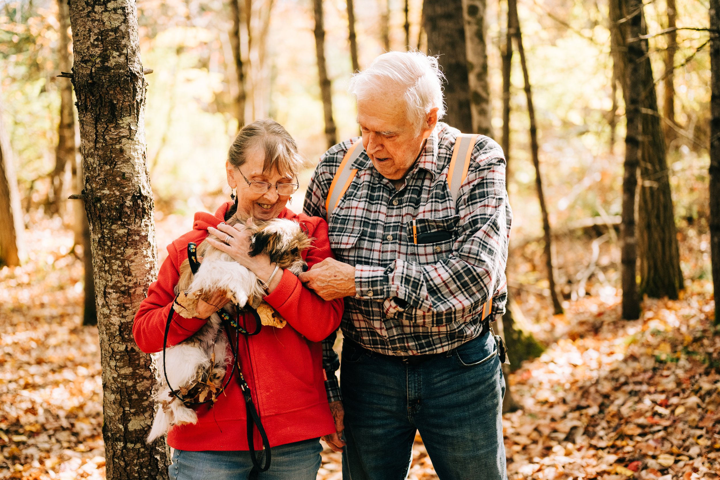 An elderly couple in a forest during autumn, with the woman holding a small dog and both smiling at it.
