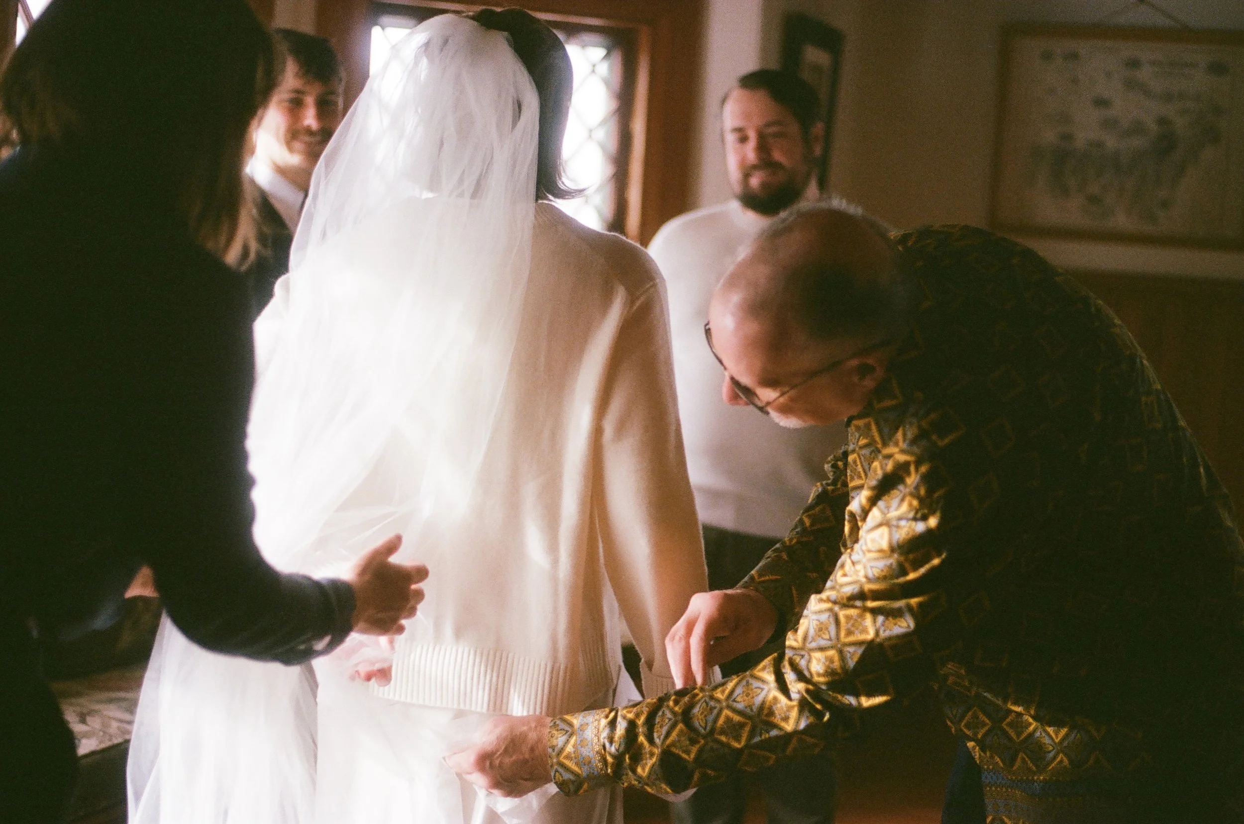 A bride getting fitted for her wedding dress with several people assisting her, including a man in a patterned shirt adjusting the dress and others observing in a warmly lit room.