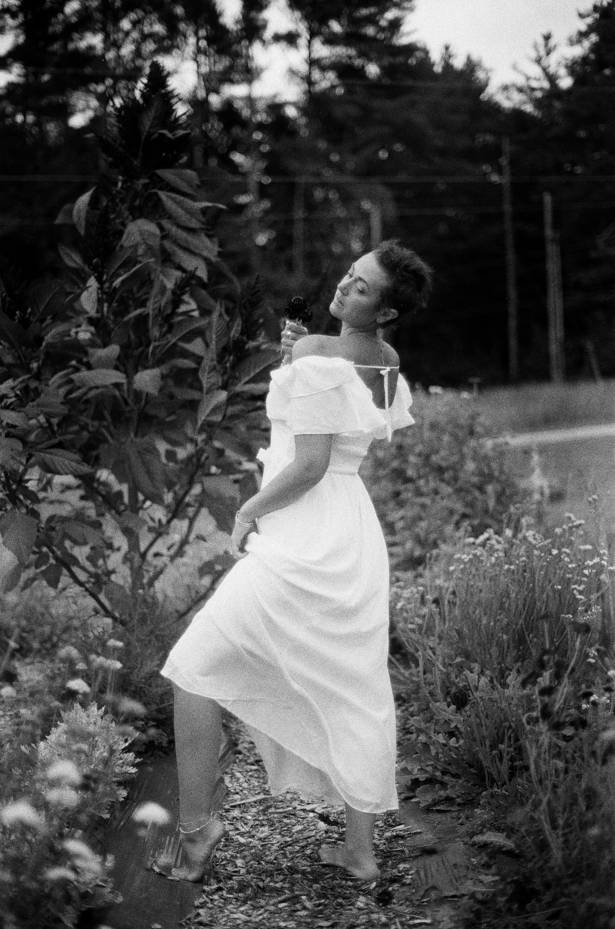 A woman in a long, light-colored dress holding an ice cream cone, standing outdoors beside a bush, with trees and power lines in the background.