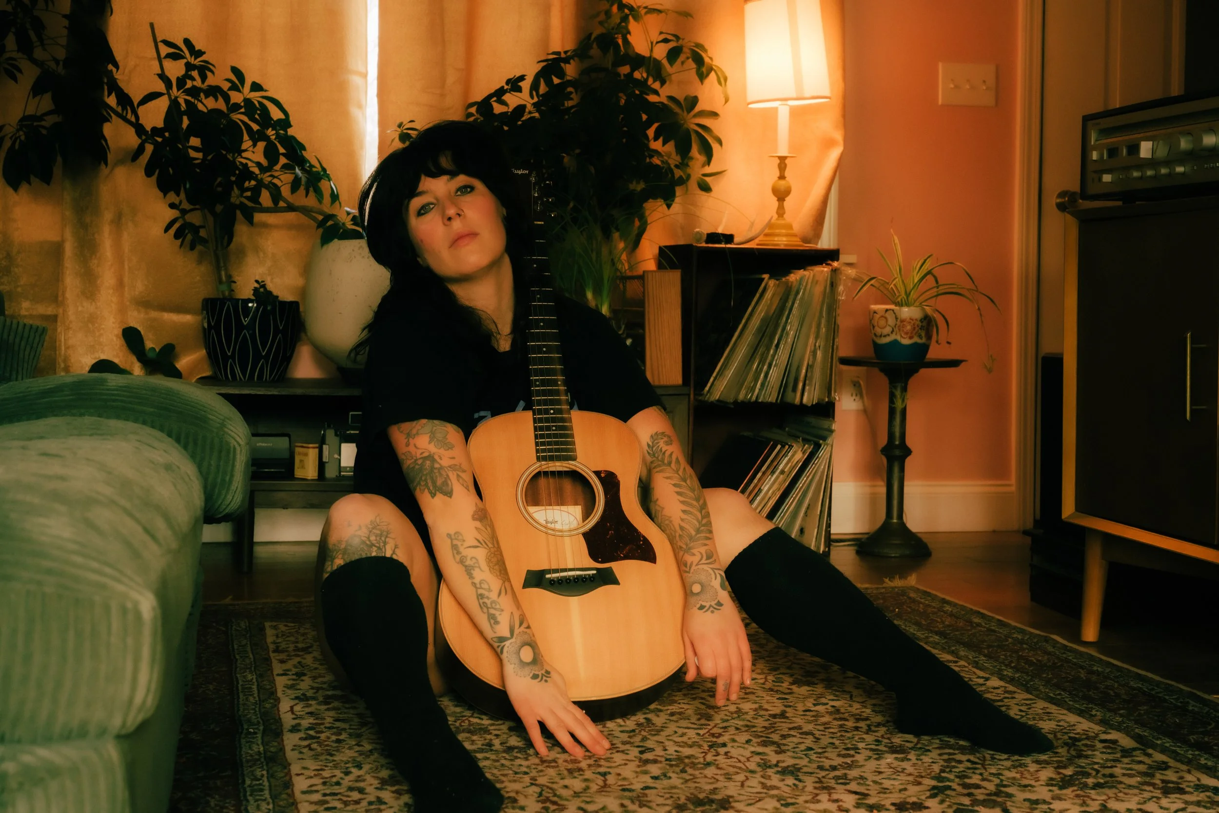 A woman with tattoos sitting on a rug in a living room, holding an acoustic guitar against her body. She has black hair and is wearing black knee-high socks and a black shirt. The room is warmly lit with a lamp, and there are plants, books, and vinta