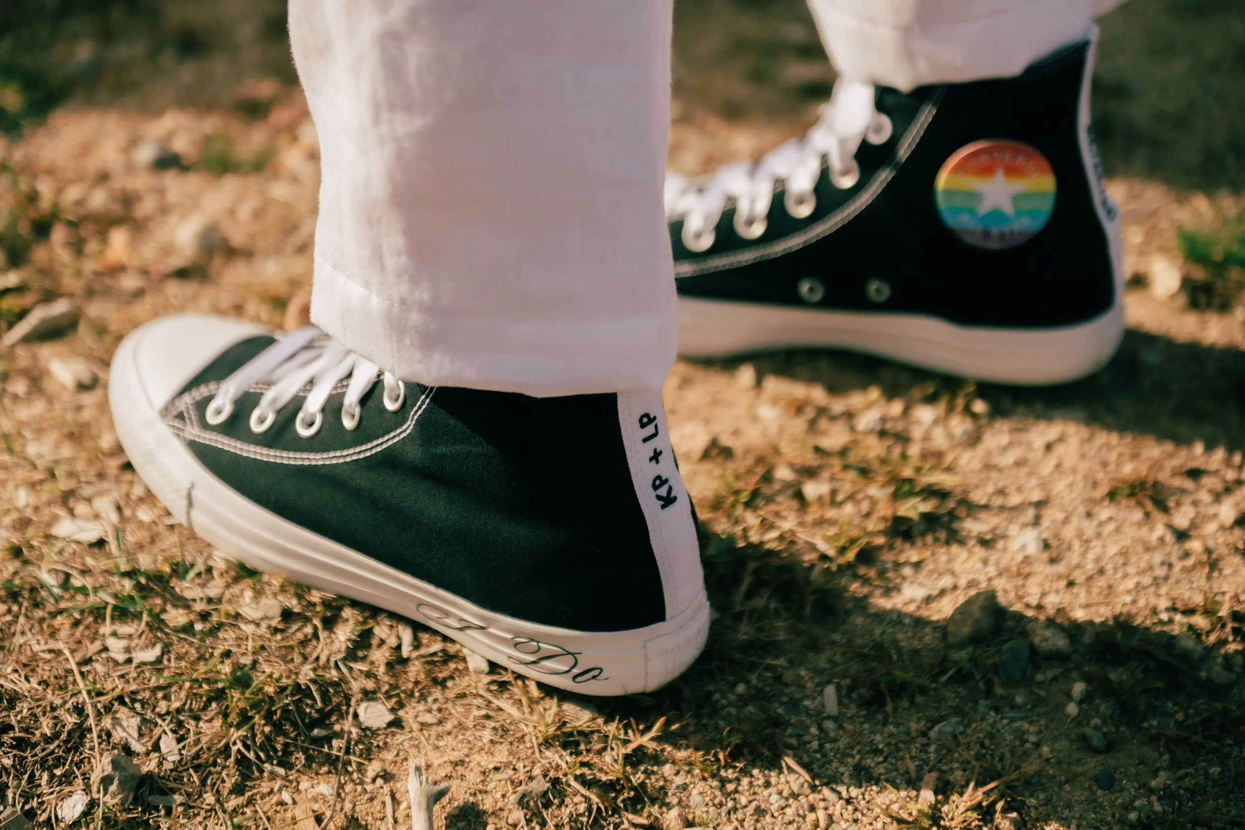 Person wearing black high-top sneakers with rainbow pride patch on the side, standing on dirt ground with small plants and rocks.