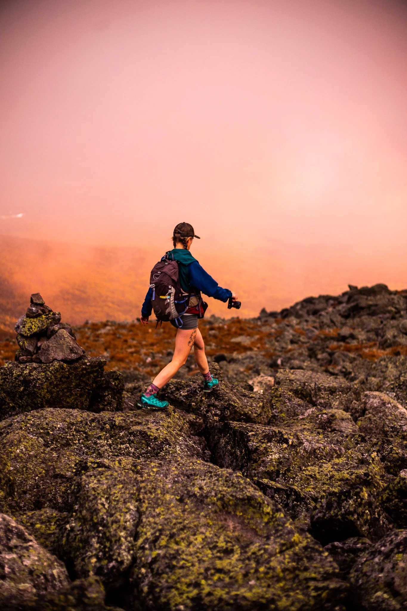 A woman hiking on rocky terrain with a backpack, holding a camera, and wearing hiking boots, a cap, and outdoor clothing, during sunset or sunrise.