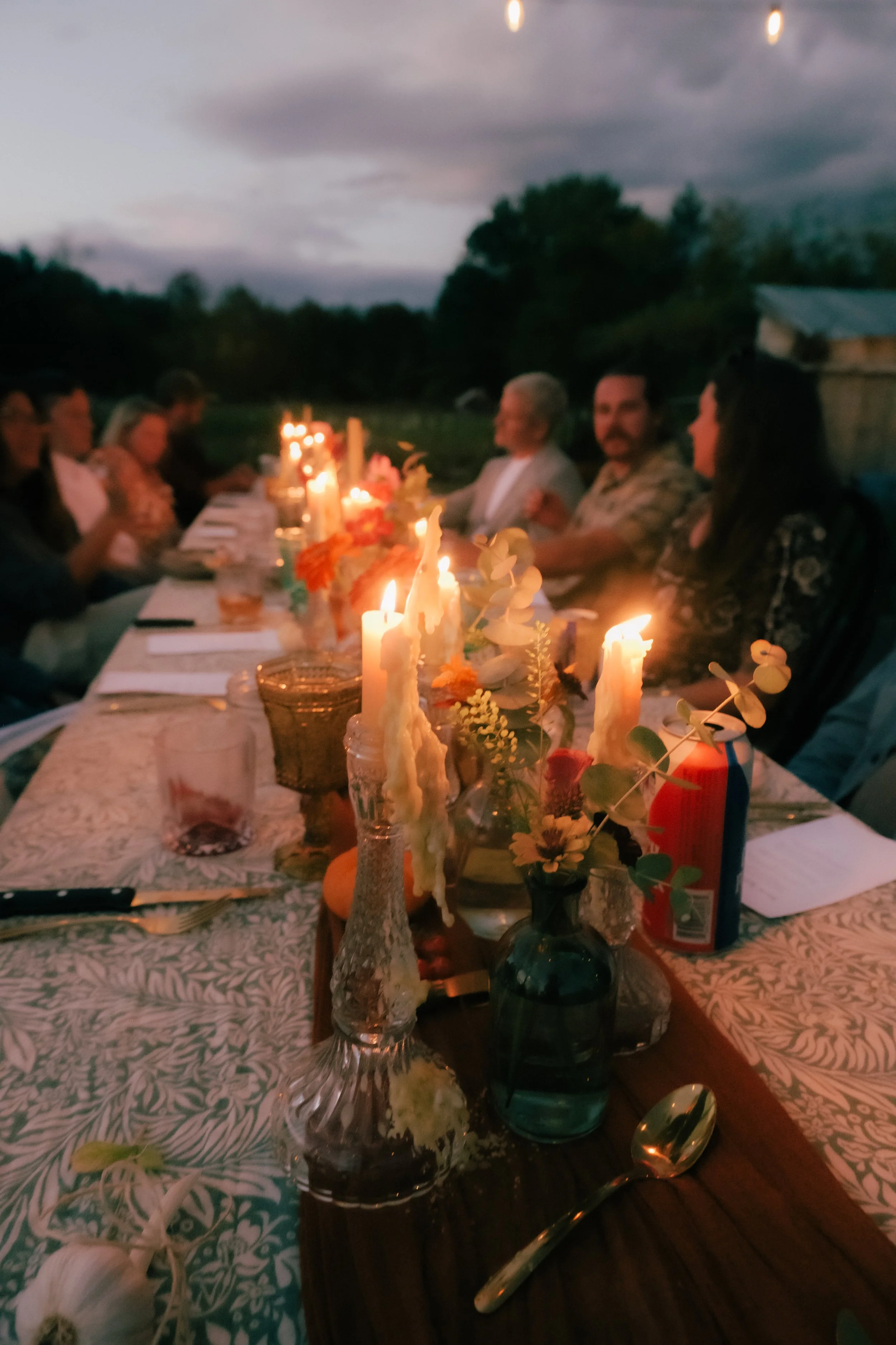 Outdoor dinner party at sunset with lit candles and guests seated at a long table.