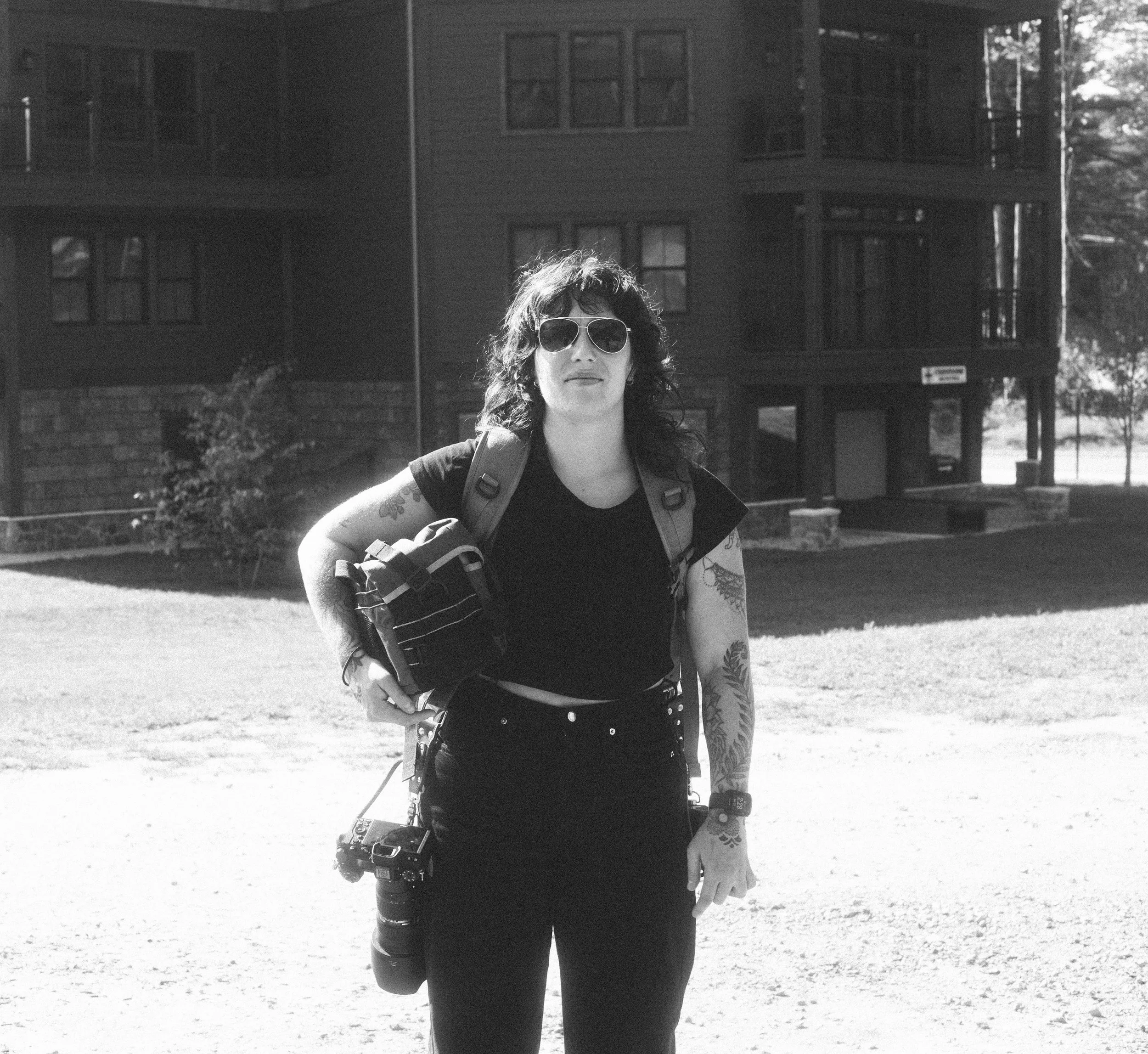 A woman with curly hair, wearing sunglasses, a black t-shirt, and black pants, standing outdoors with a backpack and a camera hanging from her waist, in front of a multi-story residential building.