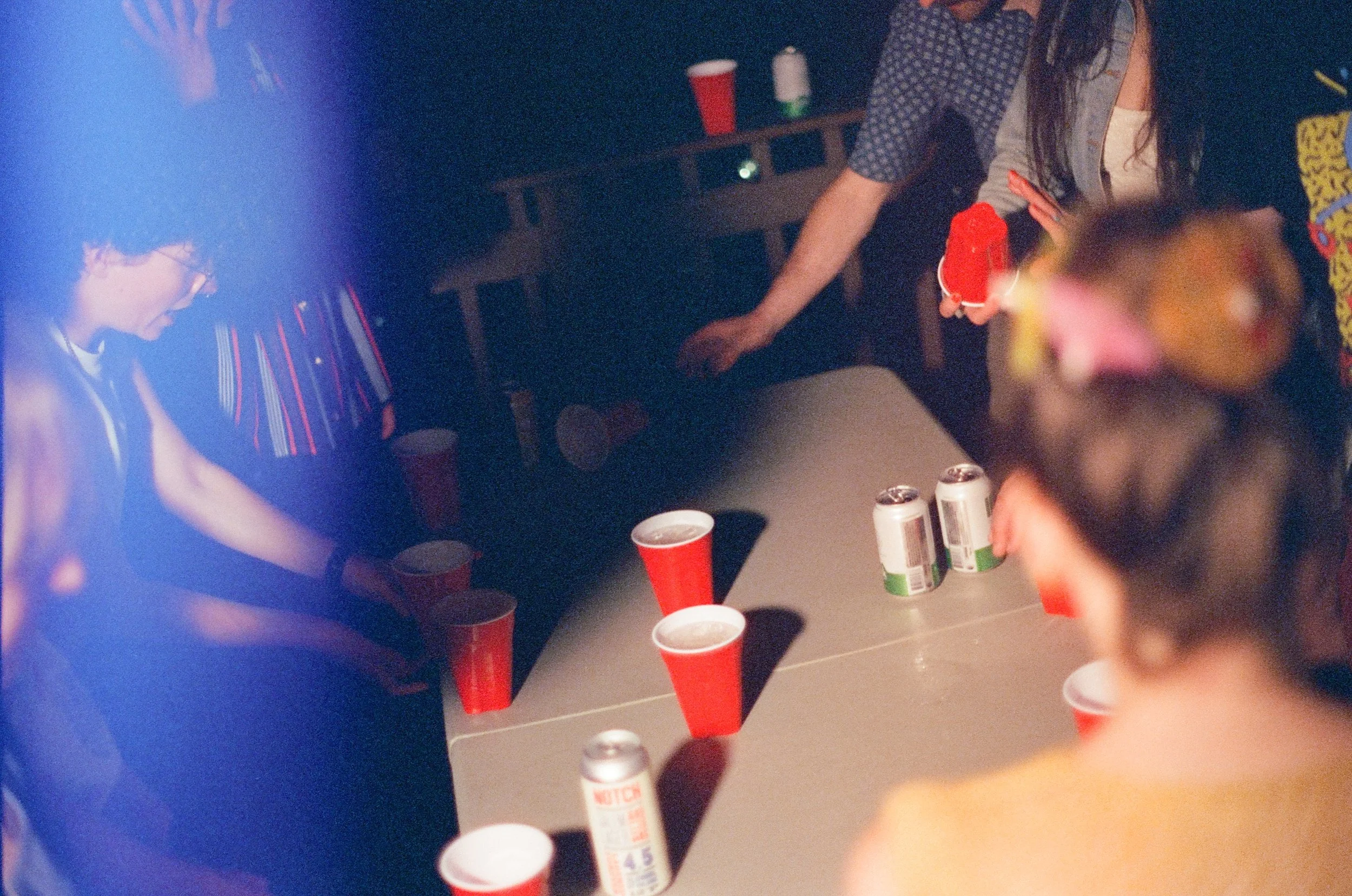 Party scene with people playing beer pong, red plastic cups, and cans on a table, dim lighting.