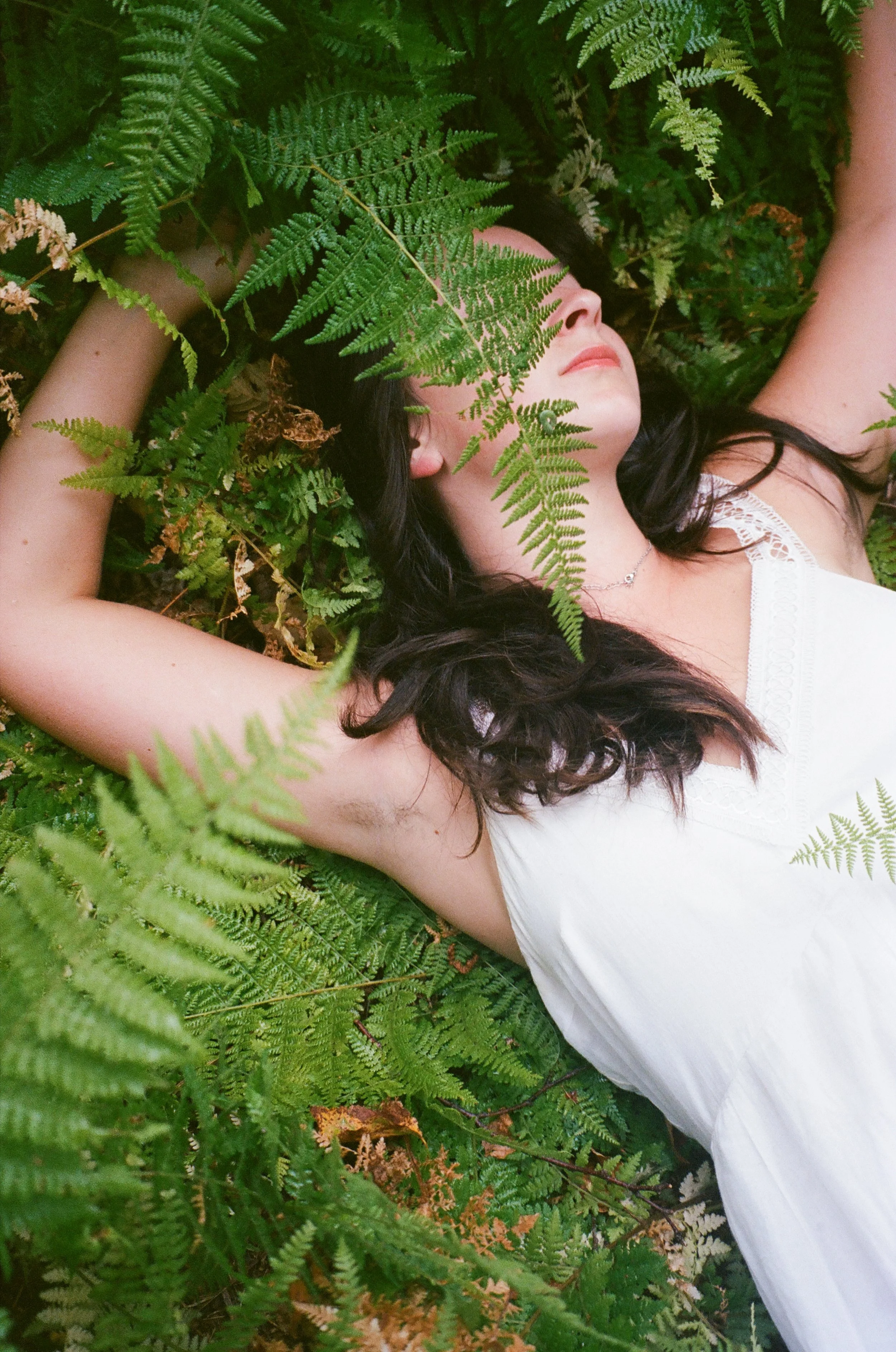A woman with dark hair lying on her back among green fern leaves, wearing a white sleeveless top.