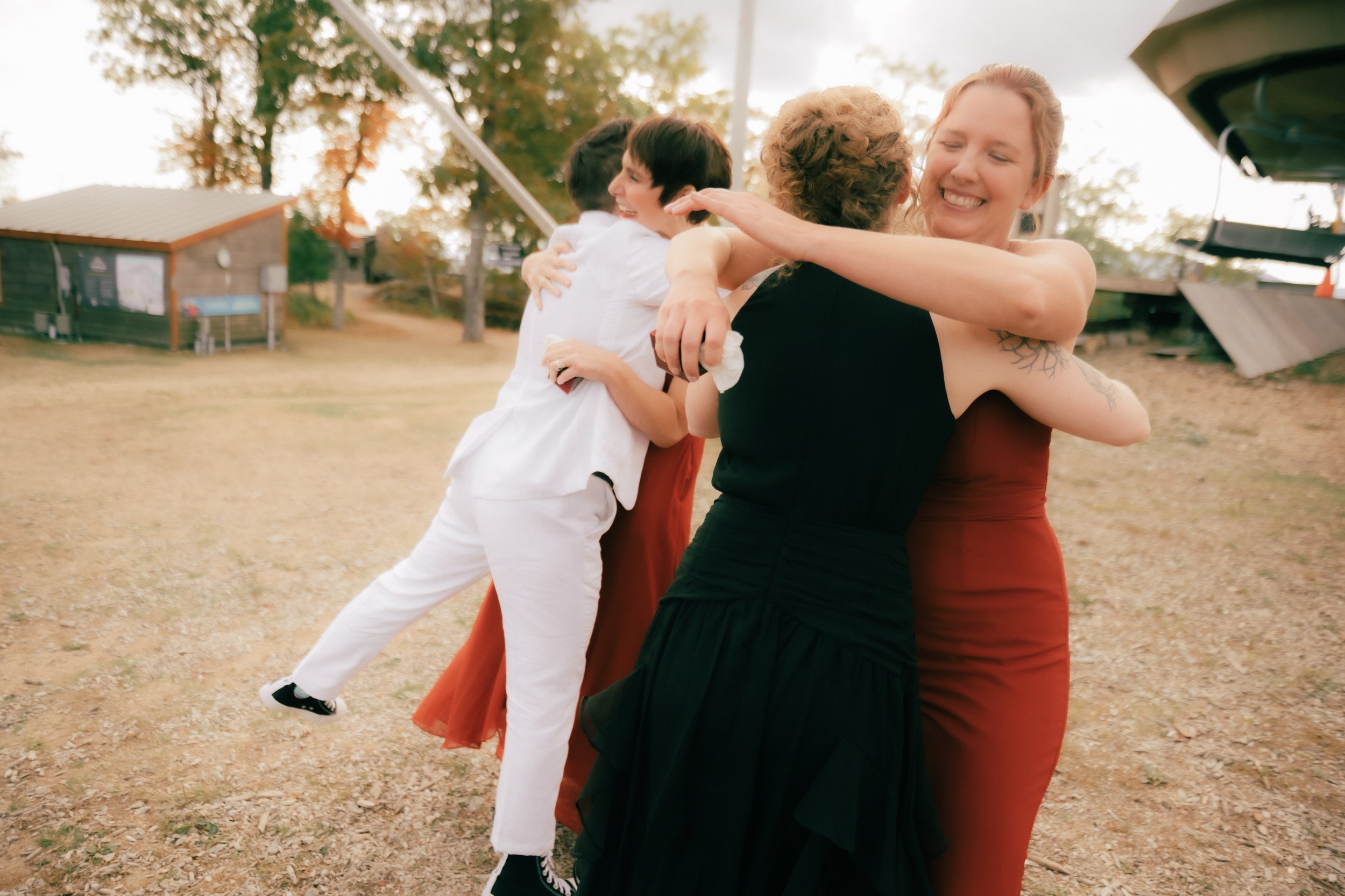Four women hugging outdoors on a sunny day, smiling, surrounded by trees and a small building.