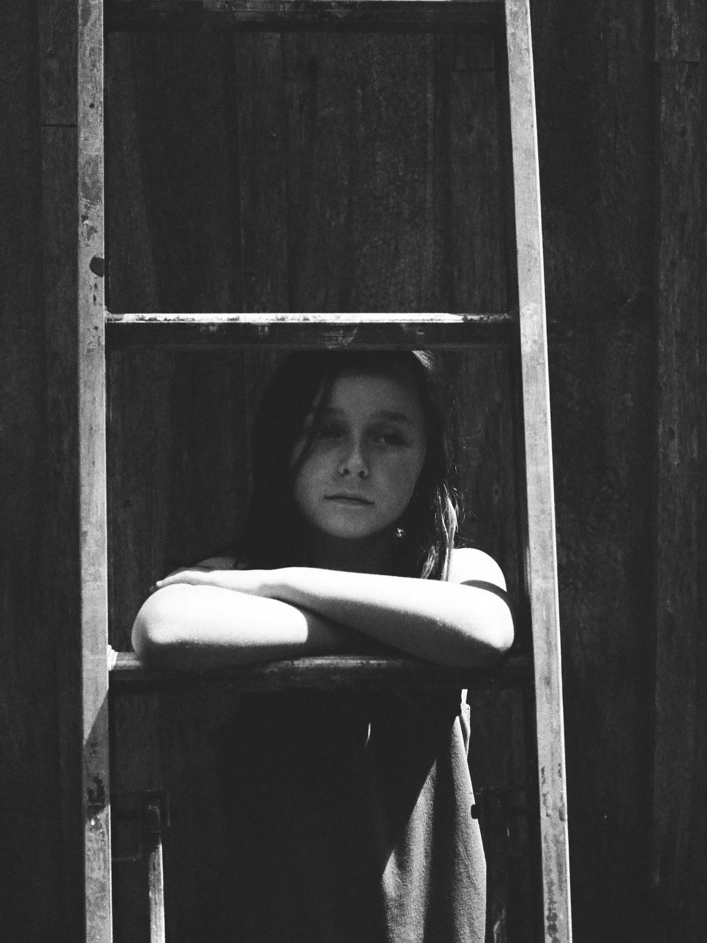 A woman with dark hair leaning on her arms, looking through a wooden window frame in black and white.