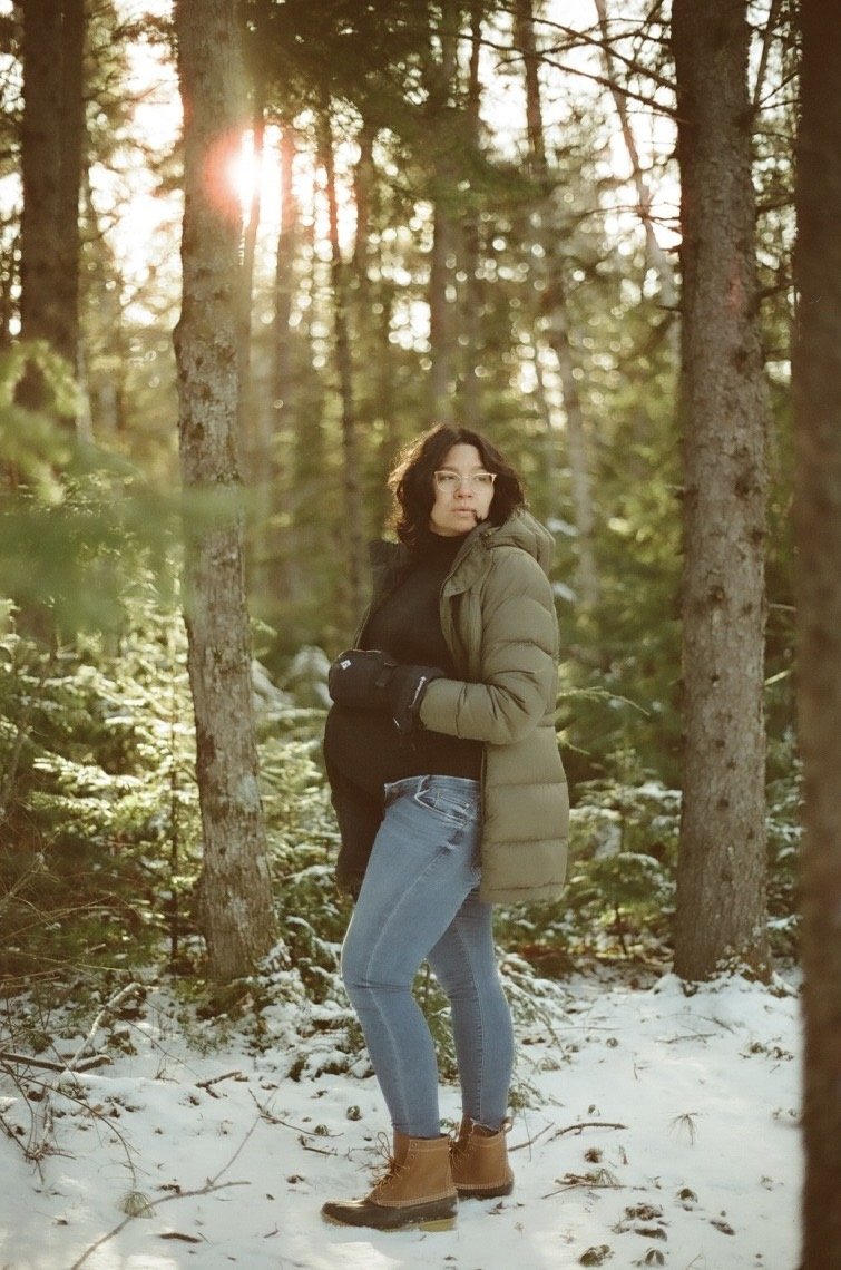A woman standing in a snowy forest with tall trees, wearing a green puffy jacket, black turtleneck, jeans, tan boots, glasses, and gloves, with sunlight filtering through the trees.
