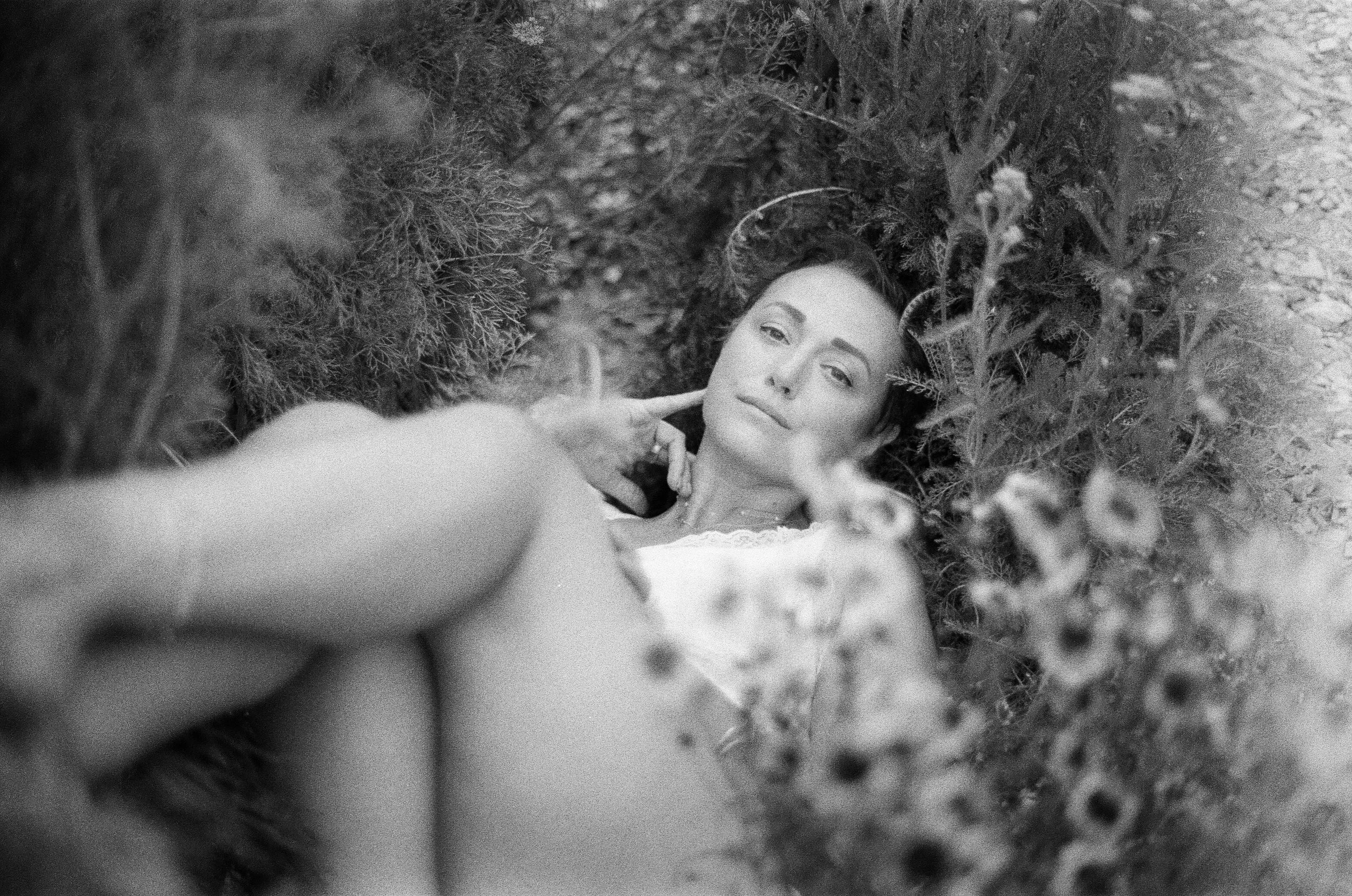 A woman lying on the ground surrounded by plants, looking at the camera with a slight smile and a finger touching her face.