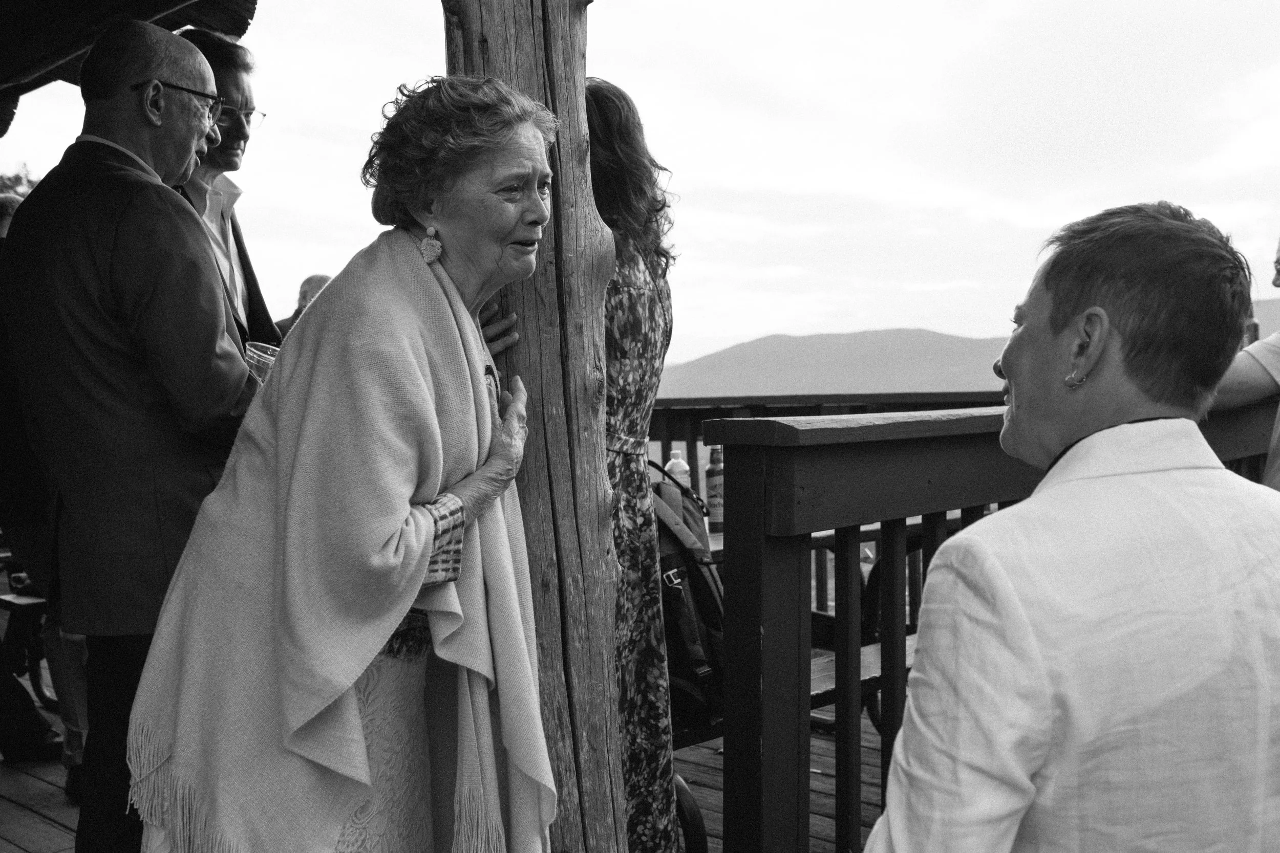 An elderly woman with short curly hair and earrings is speaking to a woman and man in a white jacket on a wooden balcony outdoors, with several people in the background and mountains in the distance.