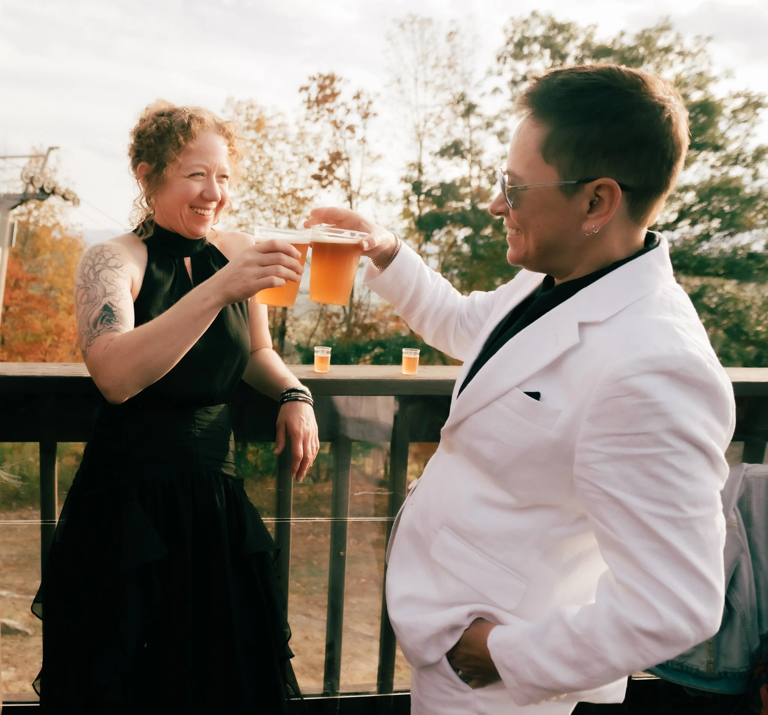 A woman and a person in a white suit cheer with glasses of beer outdoors during fall, with trees in the background.