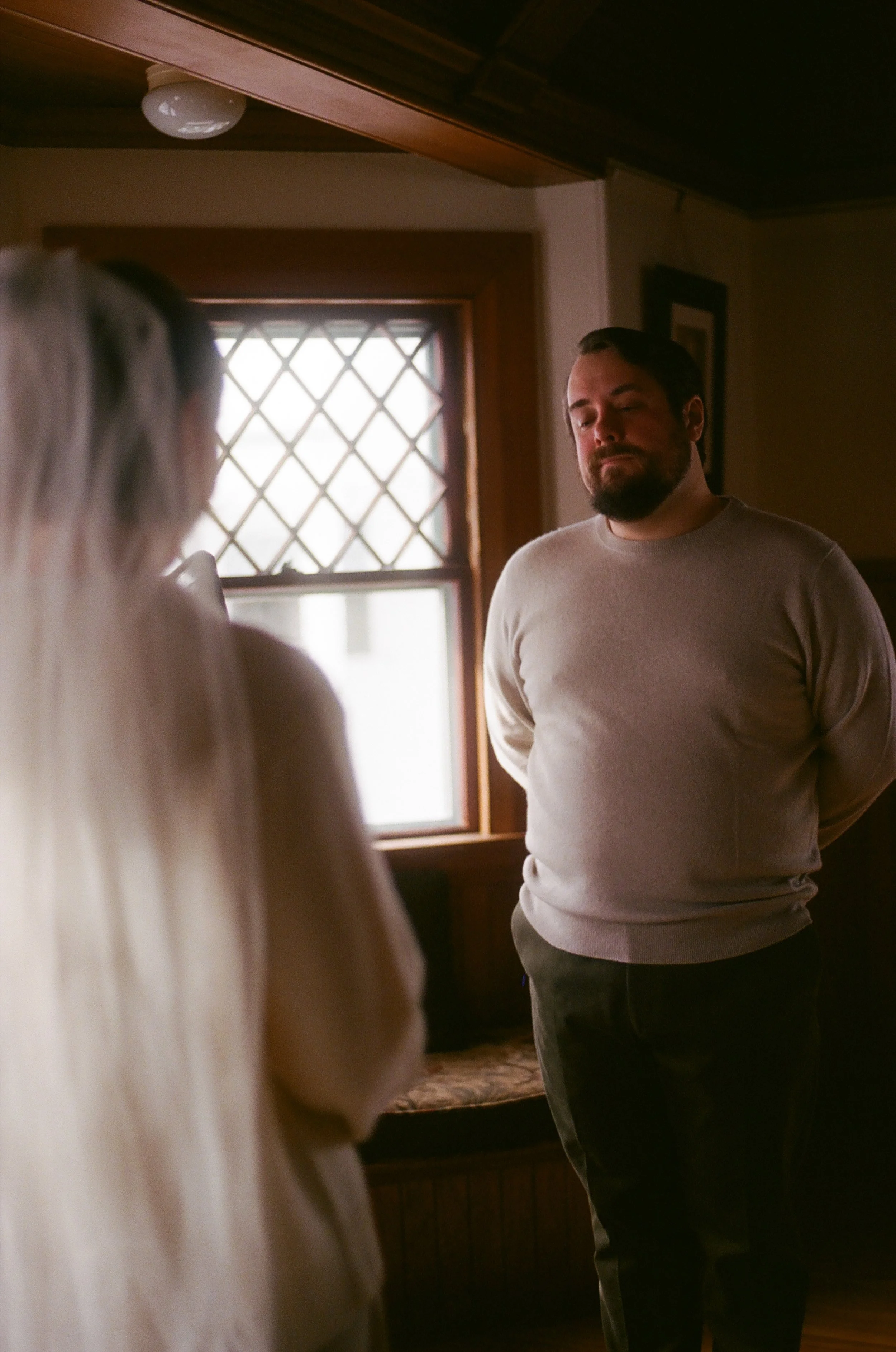 A man with a beard and long hair standing indoors, facing a woman with long blond hair, near a window with diamond-shaped panes.